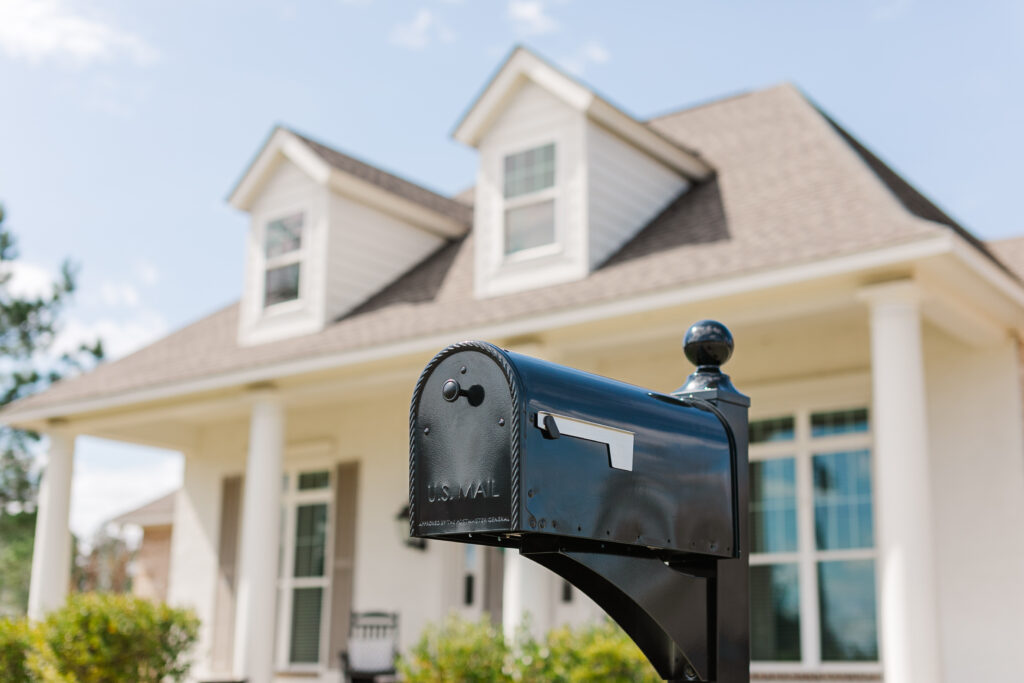 Close-up of a black U.S. Mail mailbox on a post in front of a suburban house with dormer windows, blue sky above.