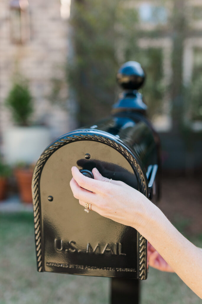 Close-up of a hand with rings dropping a letter into a black US Mail mailbox outdoors.