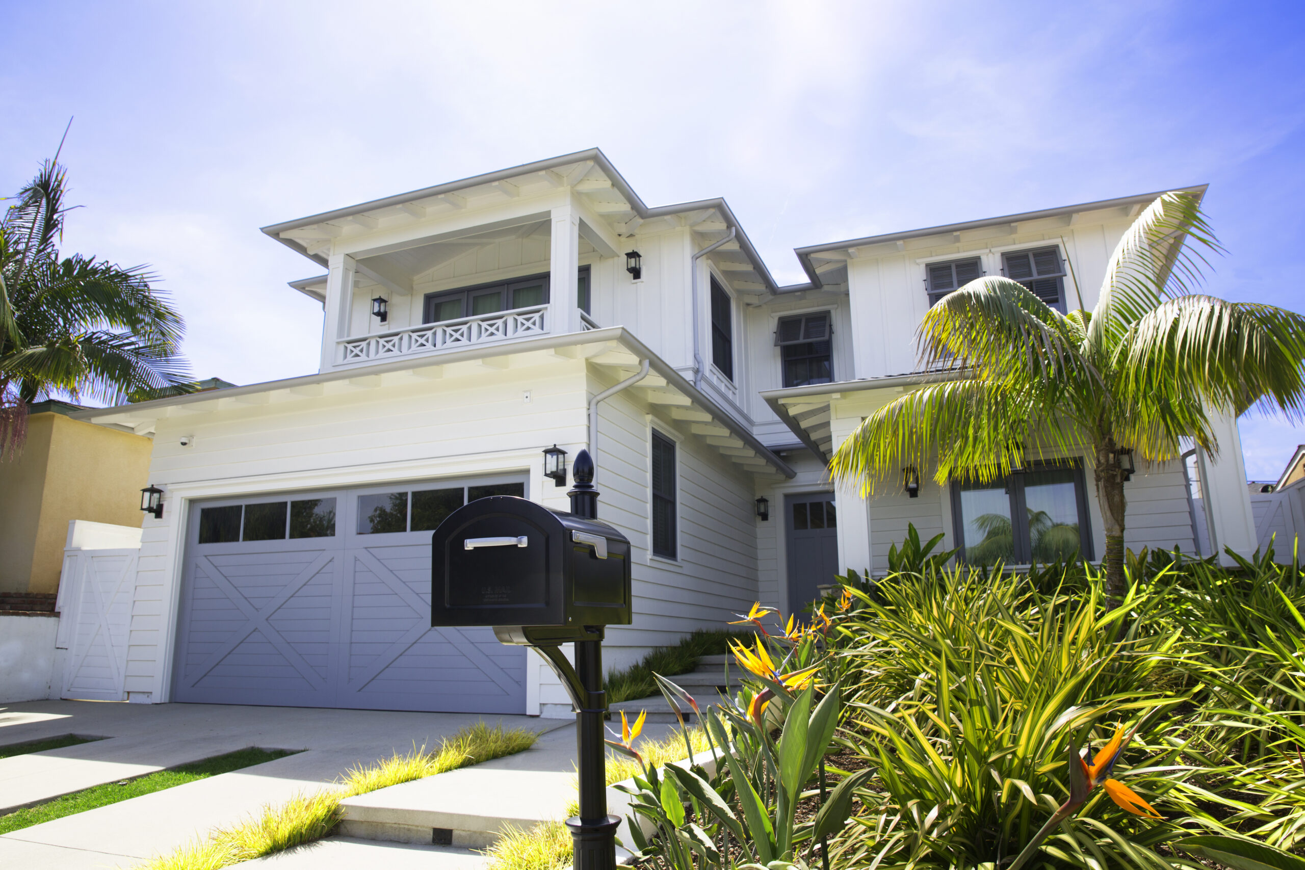 Front view of a white two-story house with a gray two-car garage, black mailbox in the foreground, and tropical landscaping including palm trees.