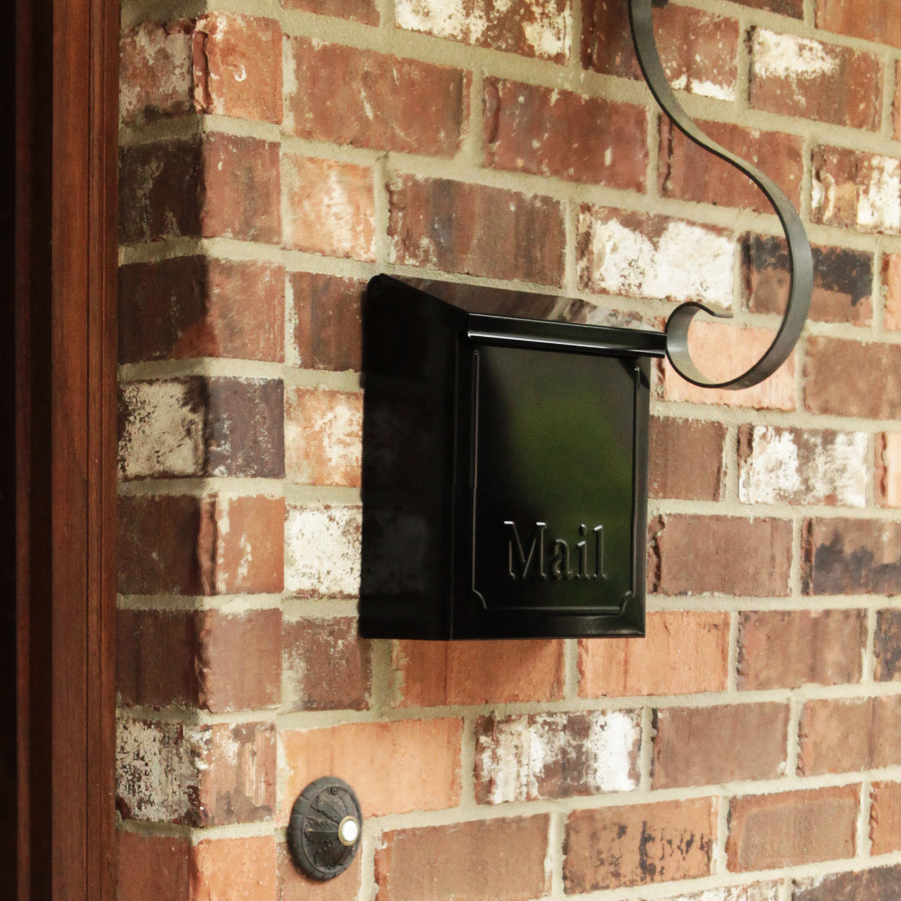 Black wall-mounted mailbox with 'Mail' embossed on the front, attached to a red brick wall with a curved metal bracket above and a doorbell below left.