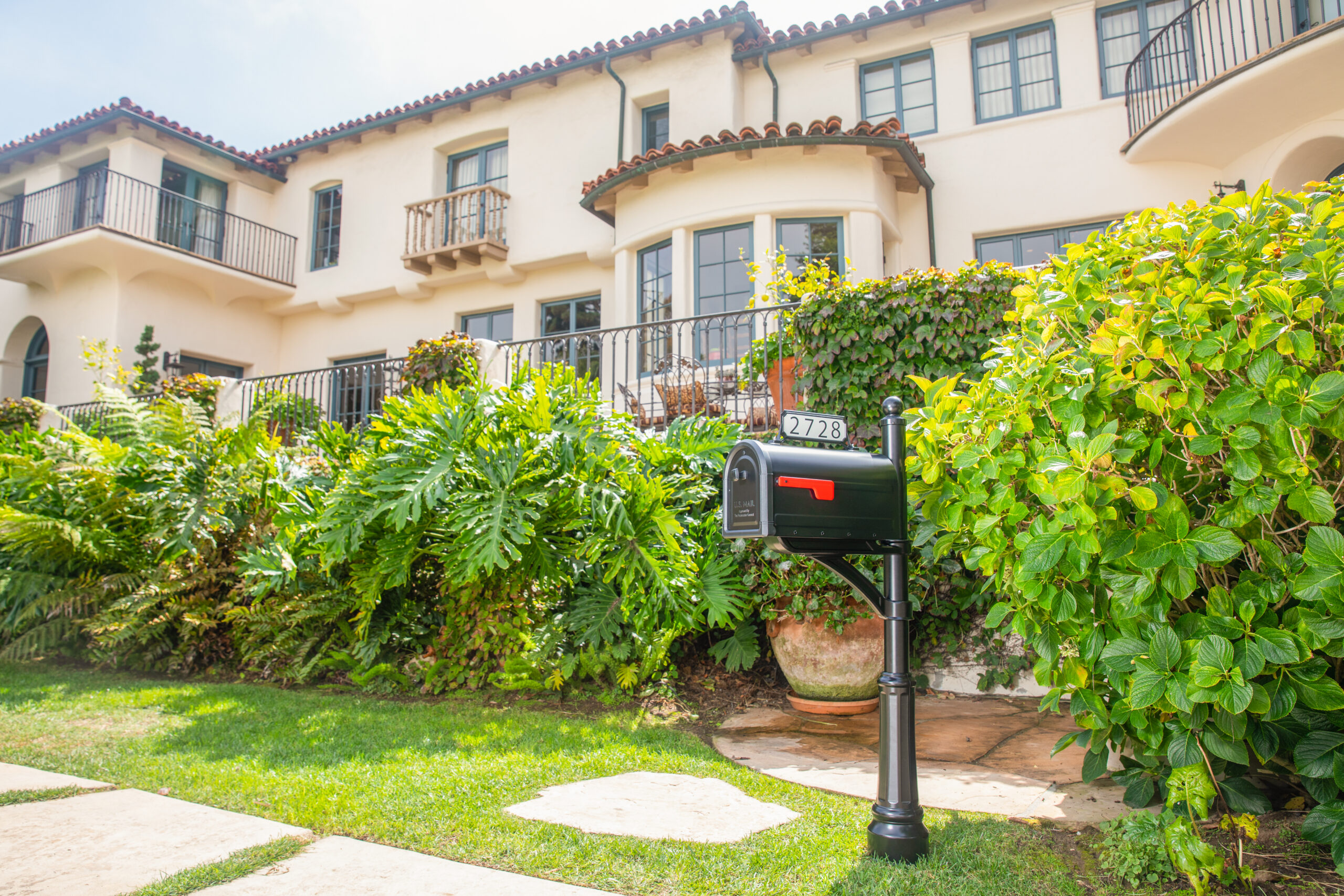 Black mailbox with a red flag on a tall black post in a sunny front yard, with a beige Mediterranean-style house and green hedges in the background. Address 2728 is visible on the mailbox.