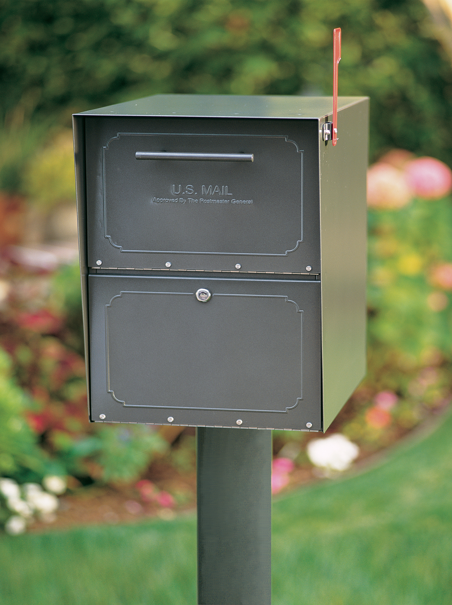 Green, two-slot U.S. Mailbox on a post outdoors, with a blurred garden in the background.
