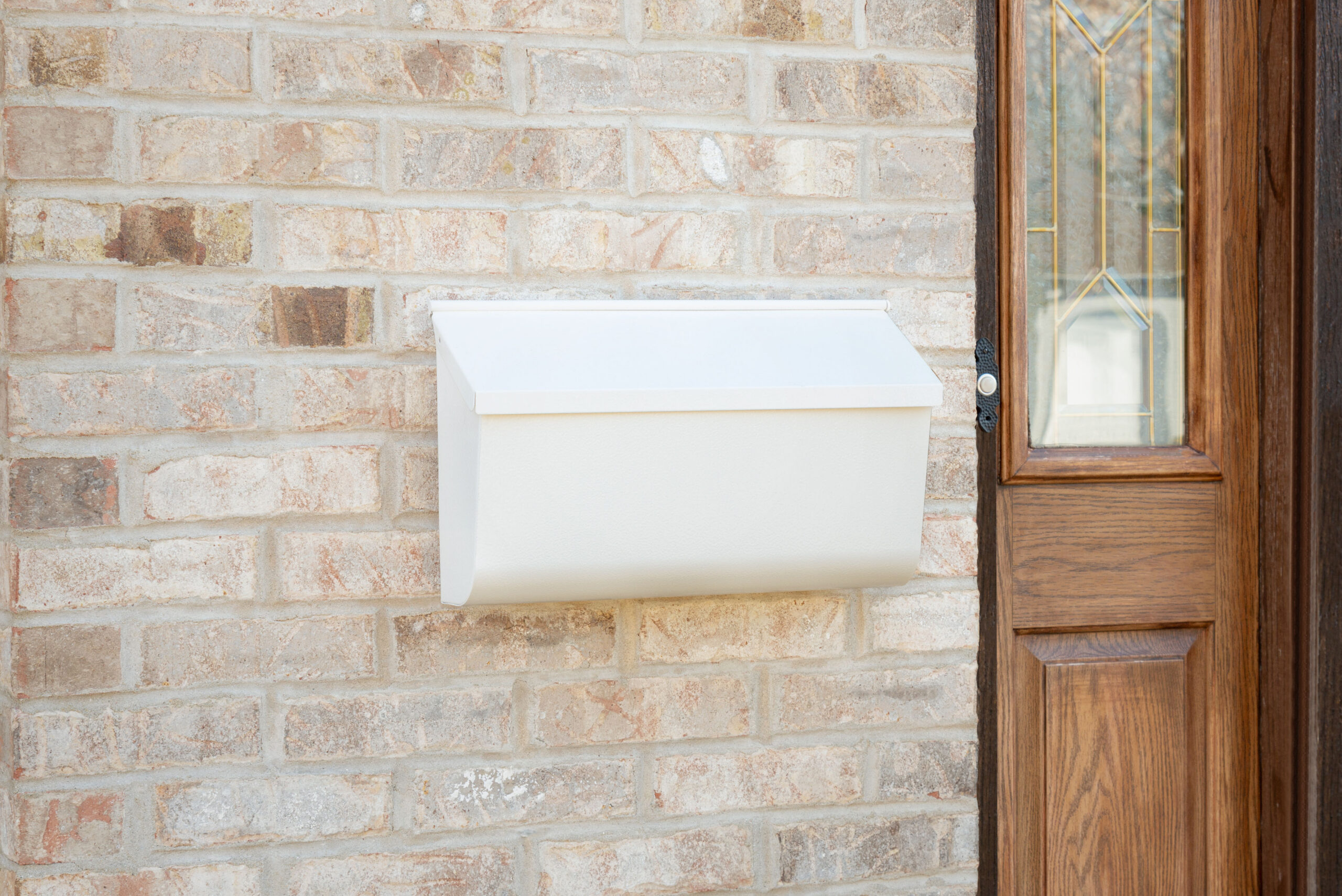 White wall-mounted mailbox on a brick exterior beside a wooden front door with a decorative glass panel area nearby.