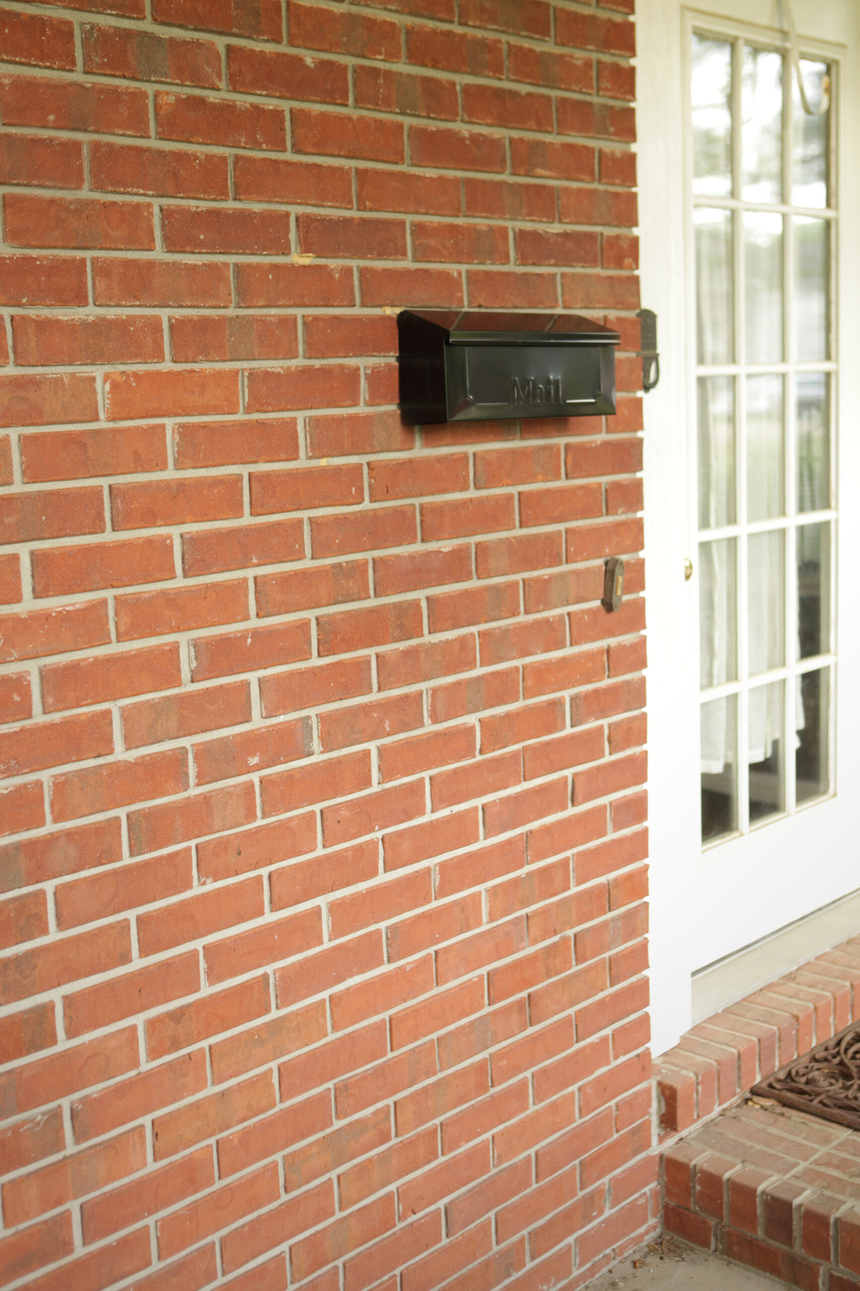 Black wall-mounted mailbox on a red brick house beside a white-framed glass door on the porch