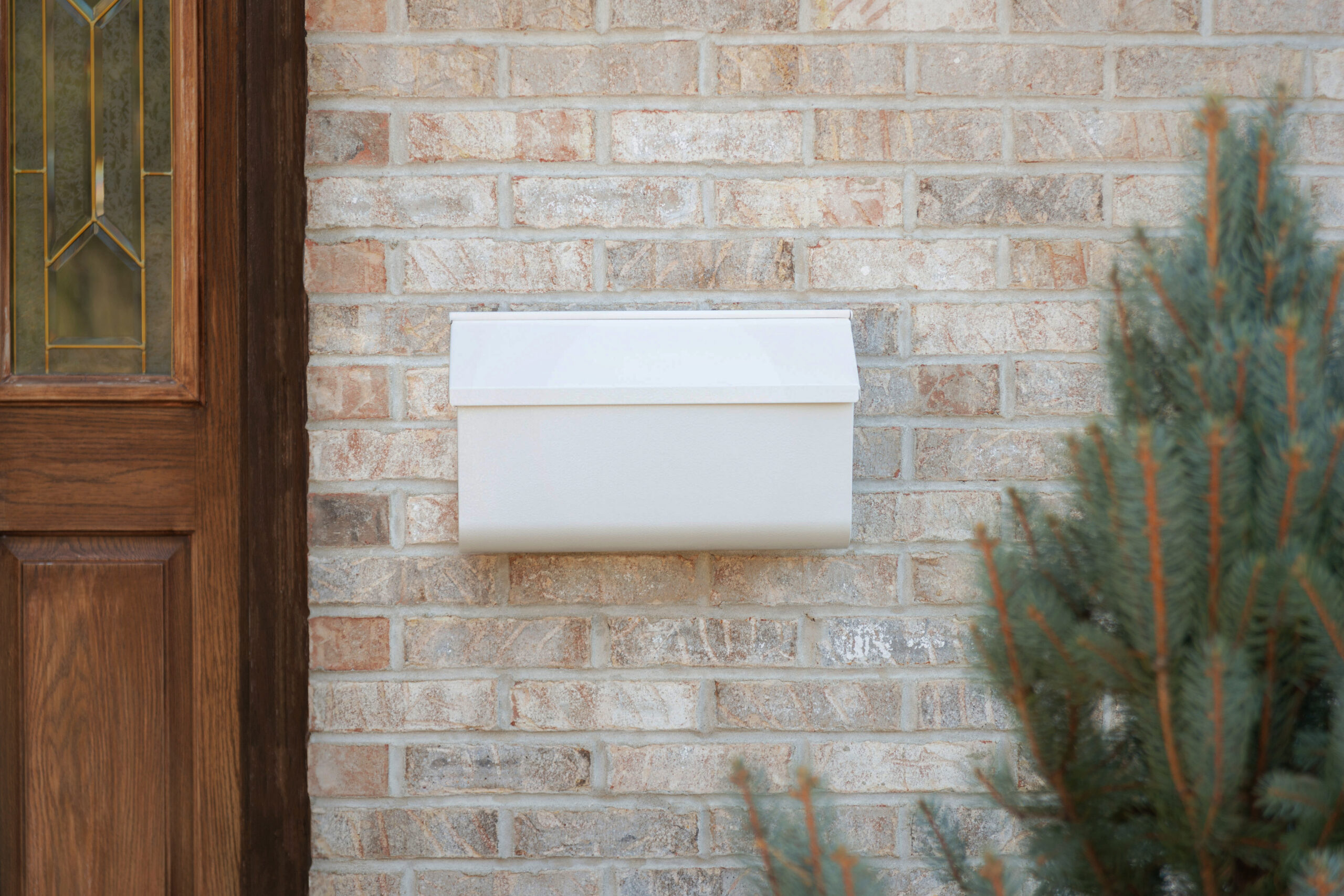 White wall-mounted mailbox on a brick exterior, with a wooden door to the left and an evergreen shrub on the right.