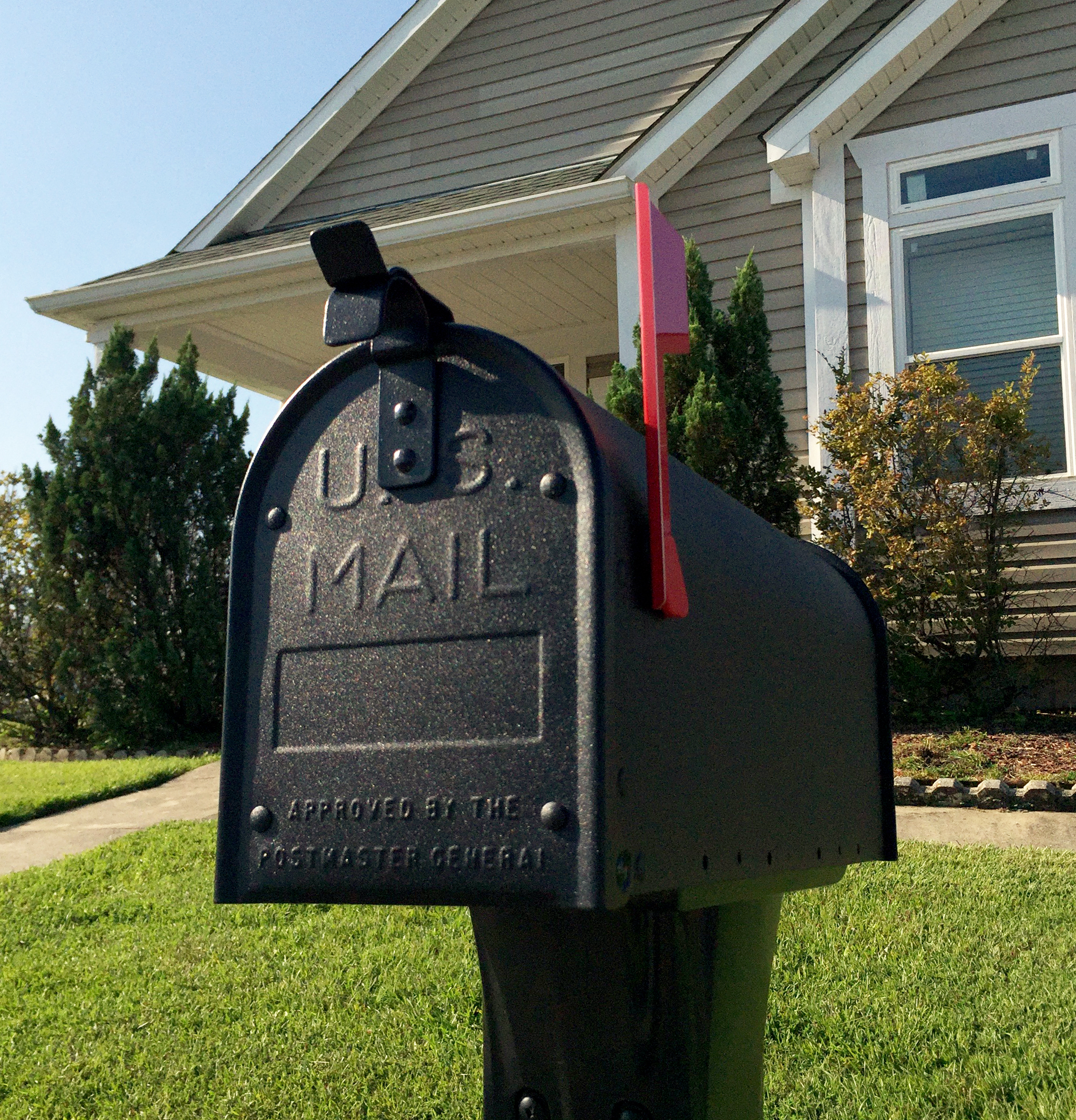 Black USPS mailbox with the red flag raised, in a front yard with a house in the background.