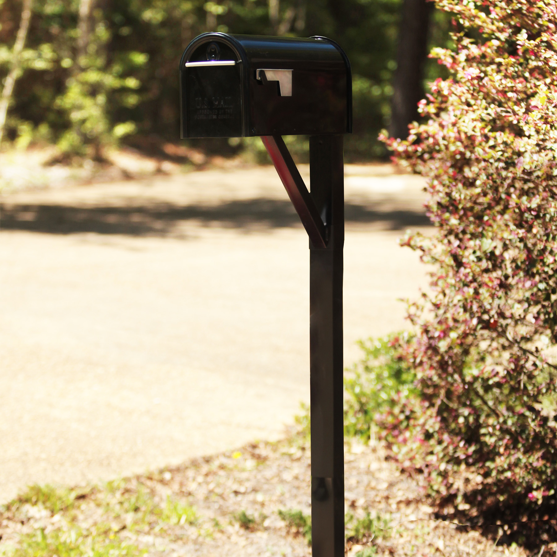 Black mailbox on a metal post beside a flowering shrub, with a red flag and a sunlit yard in the background.