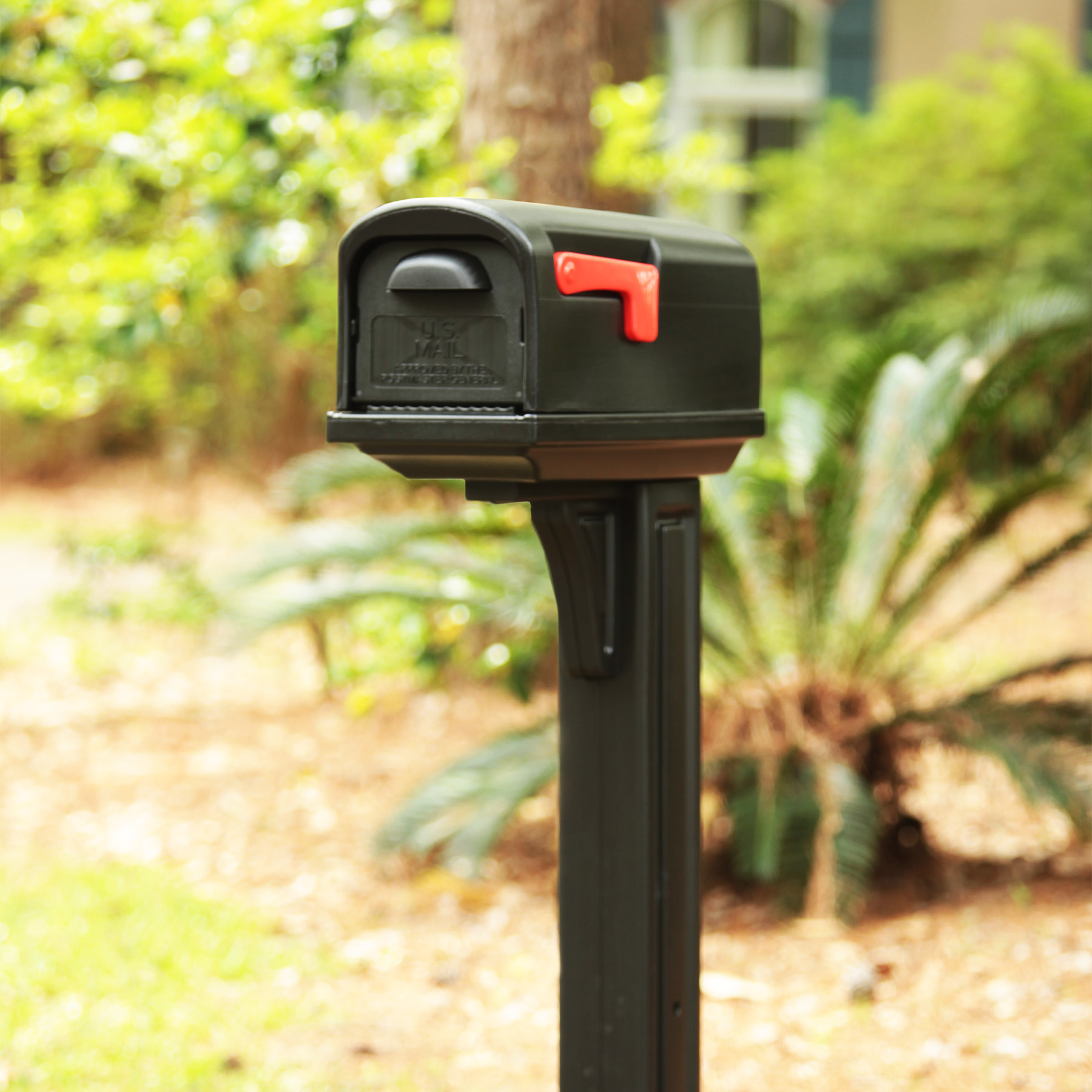 Black mailbox with a red flag mounted on a dark post in a sunny front yard garden setting