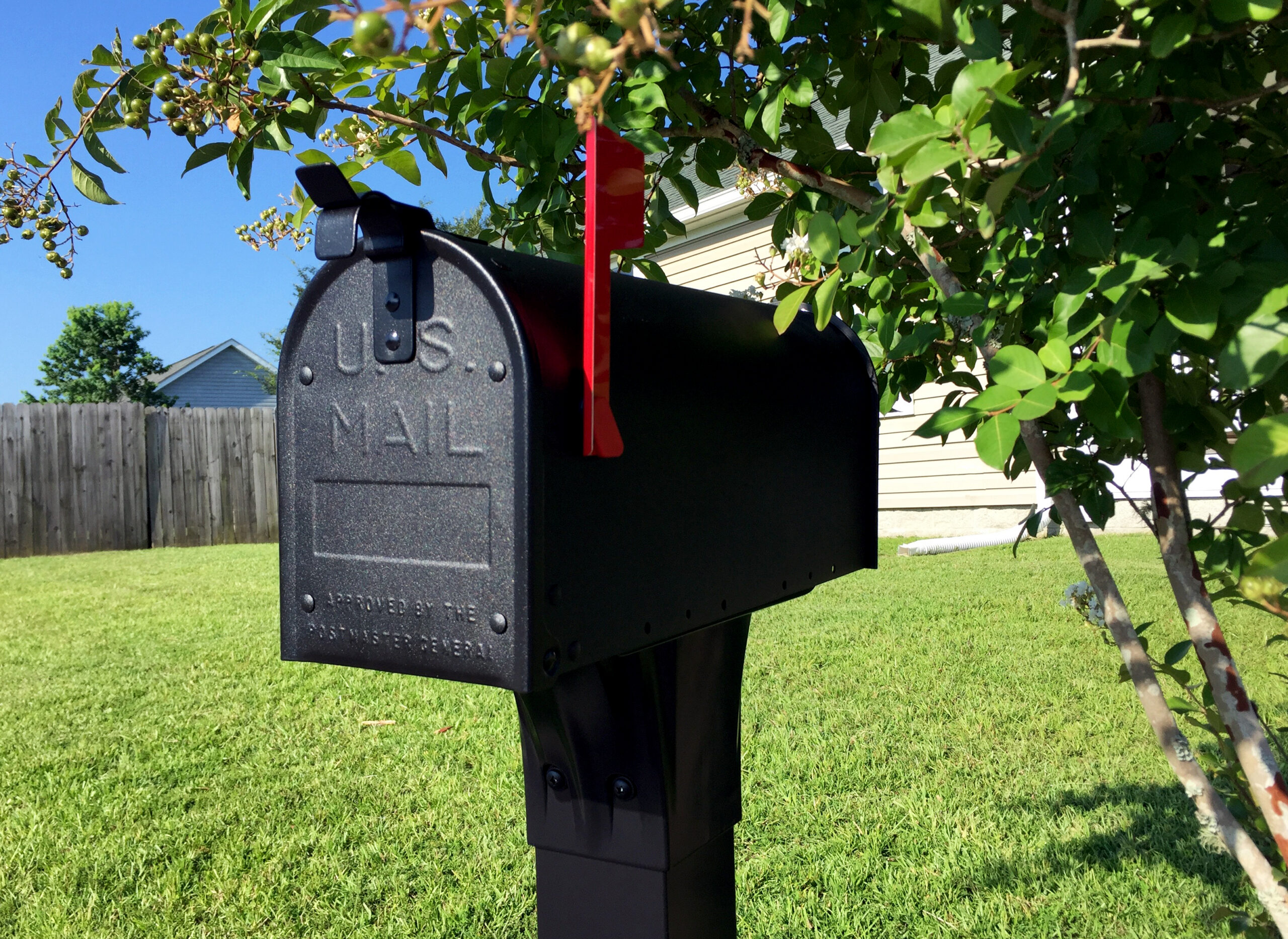 Black curbside mailbox on a post with the red flag raised, in a sunny yard with a tree and blue sky nearby.