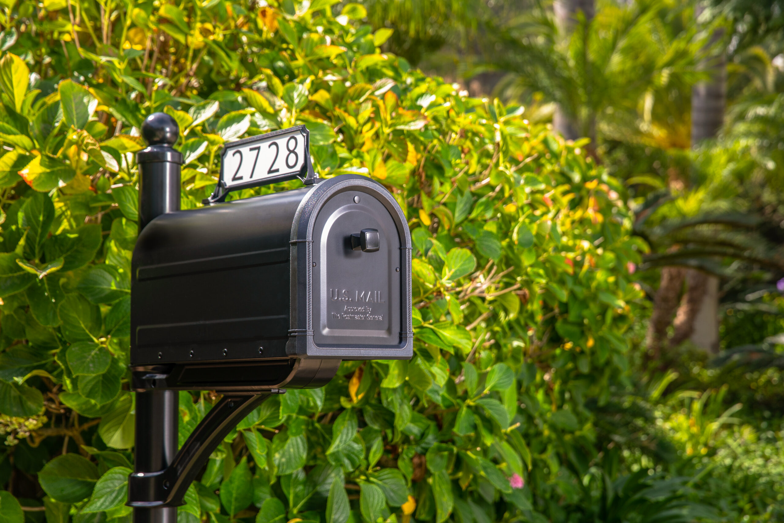 Black USPS mailbox on a post with the number 2728, set against lush green bushes in a residential yard.