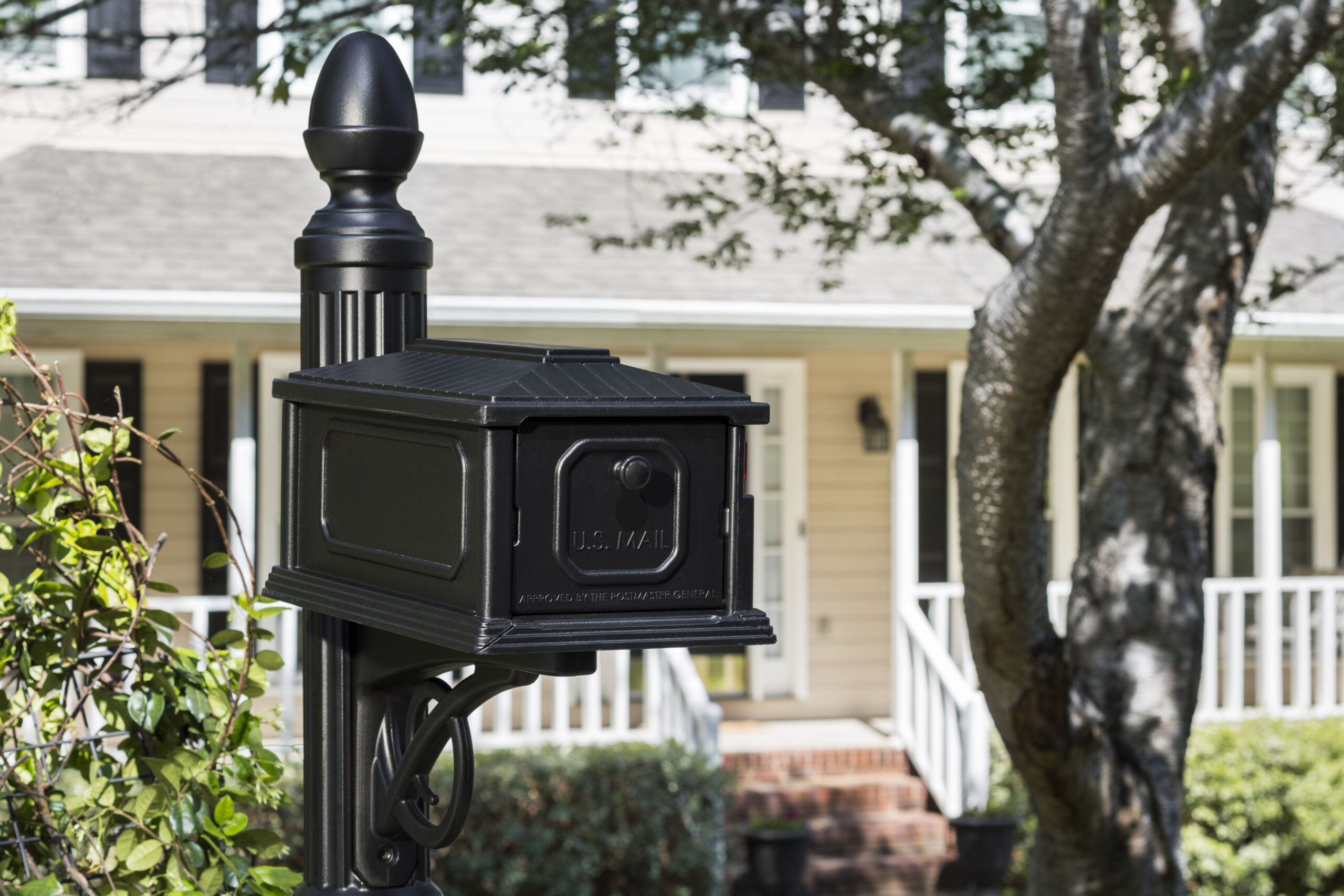 Black US mail mailbox on a post in a front-yard scene with a suburban house and white railing in the background.