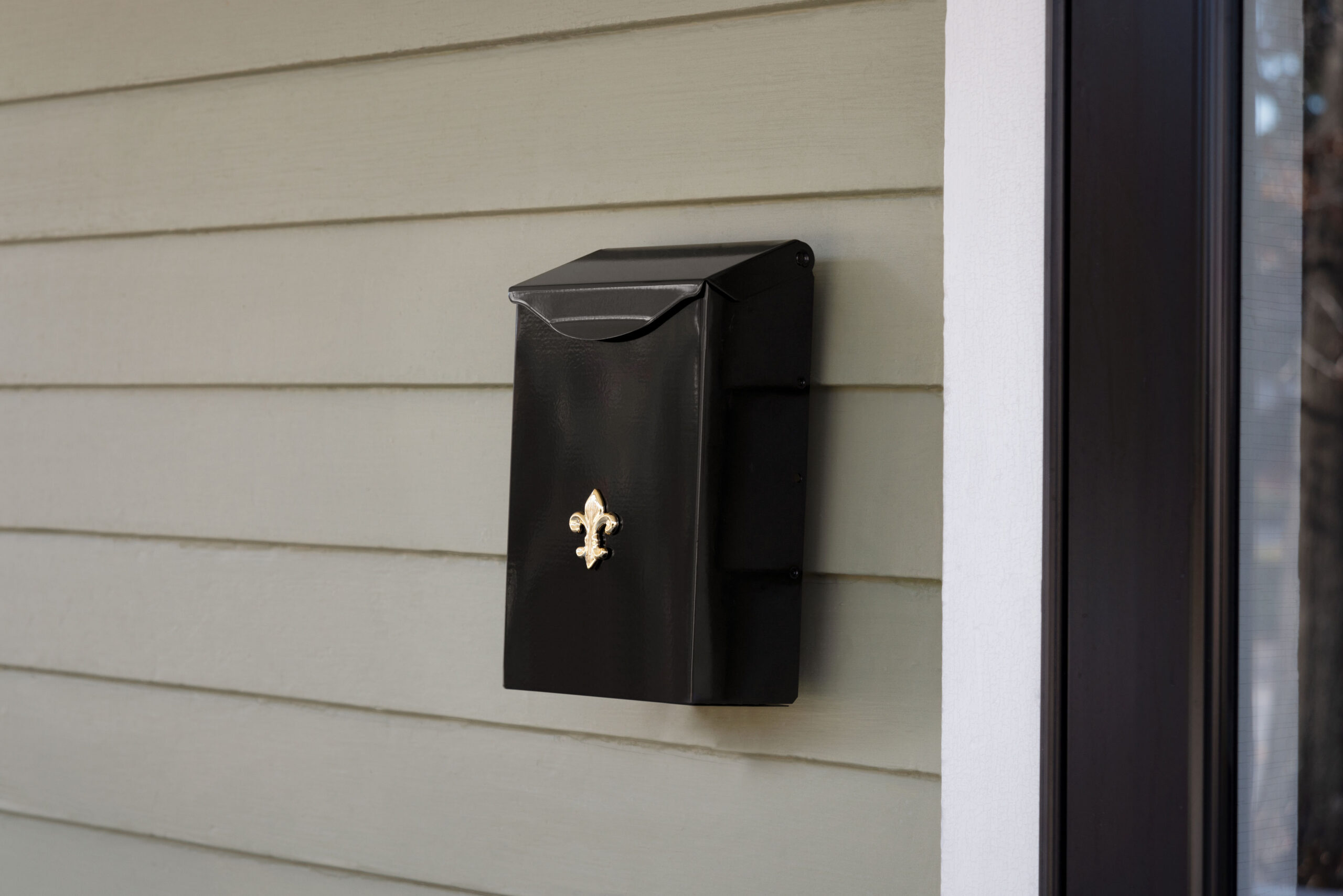 Black wall-mounted mailbox with a gold fleur-de-lis accent, attached to beige siding near a doorway.