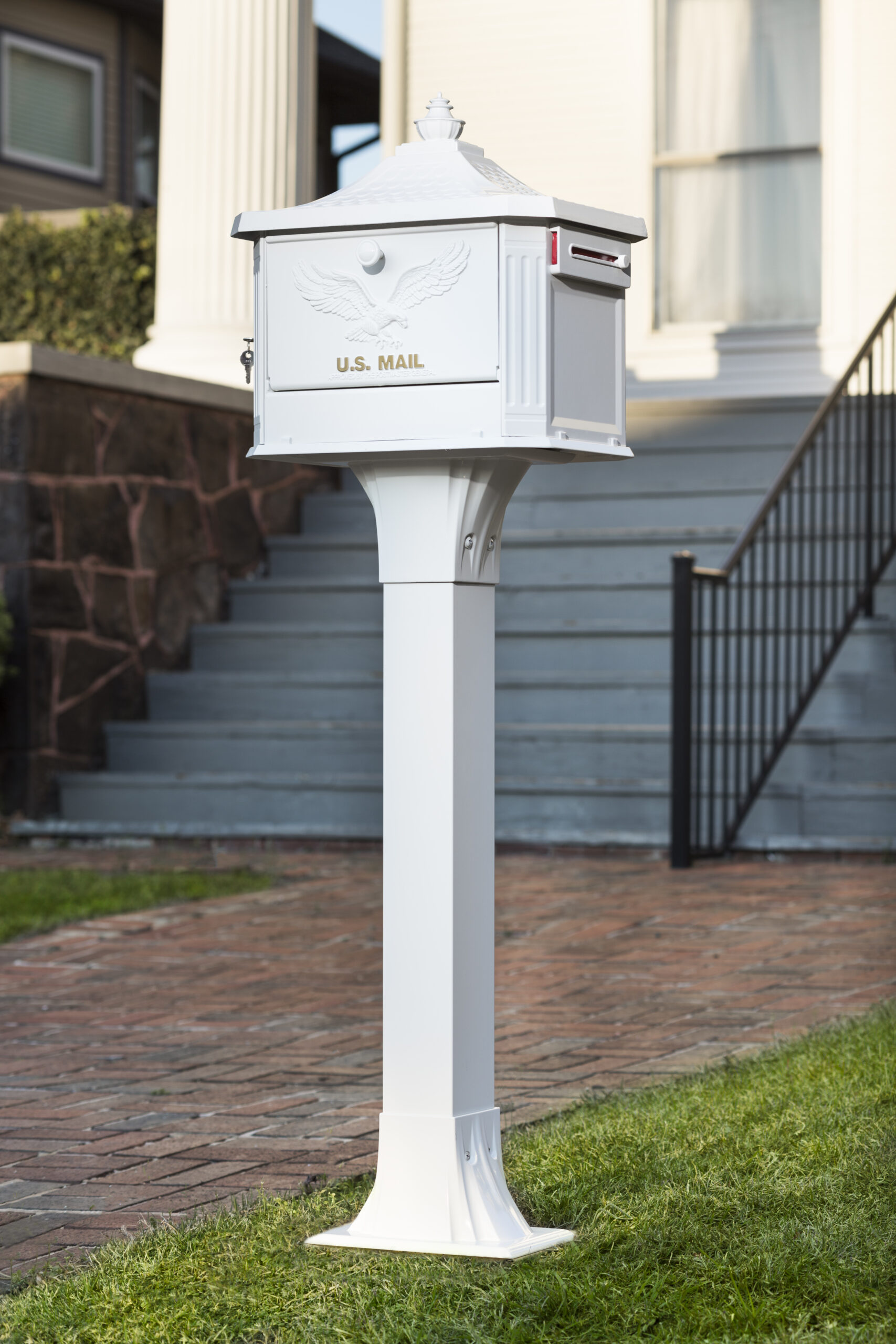 White USPS mailbox on a tall pedestal in a suburban yard by a brick walkway and stairs in the background