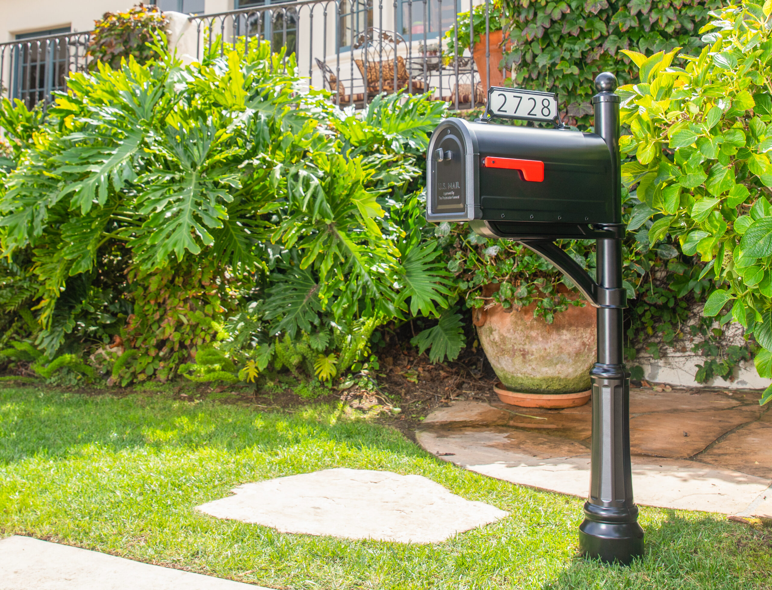 Black USPS mailbox on a tall post beside a lush green garden in bright daylight, house number 2728 visible on top of the box.