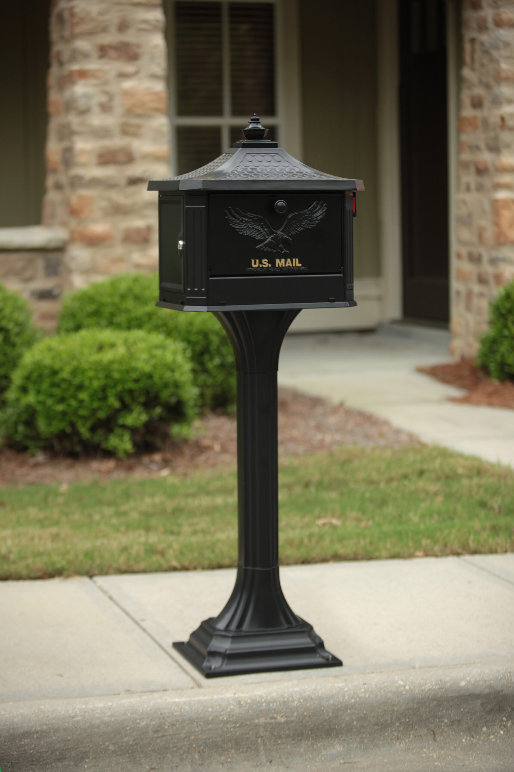 Black U.S. Mail mailbox on a tall post in a front yard with a stone house in the background.
