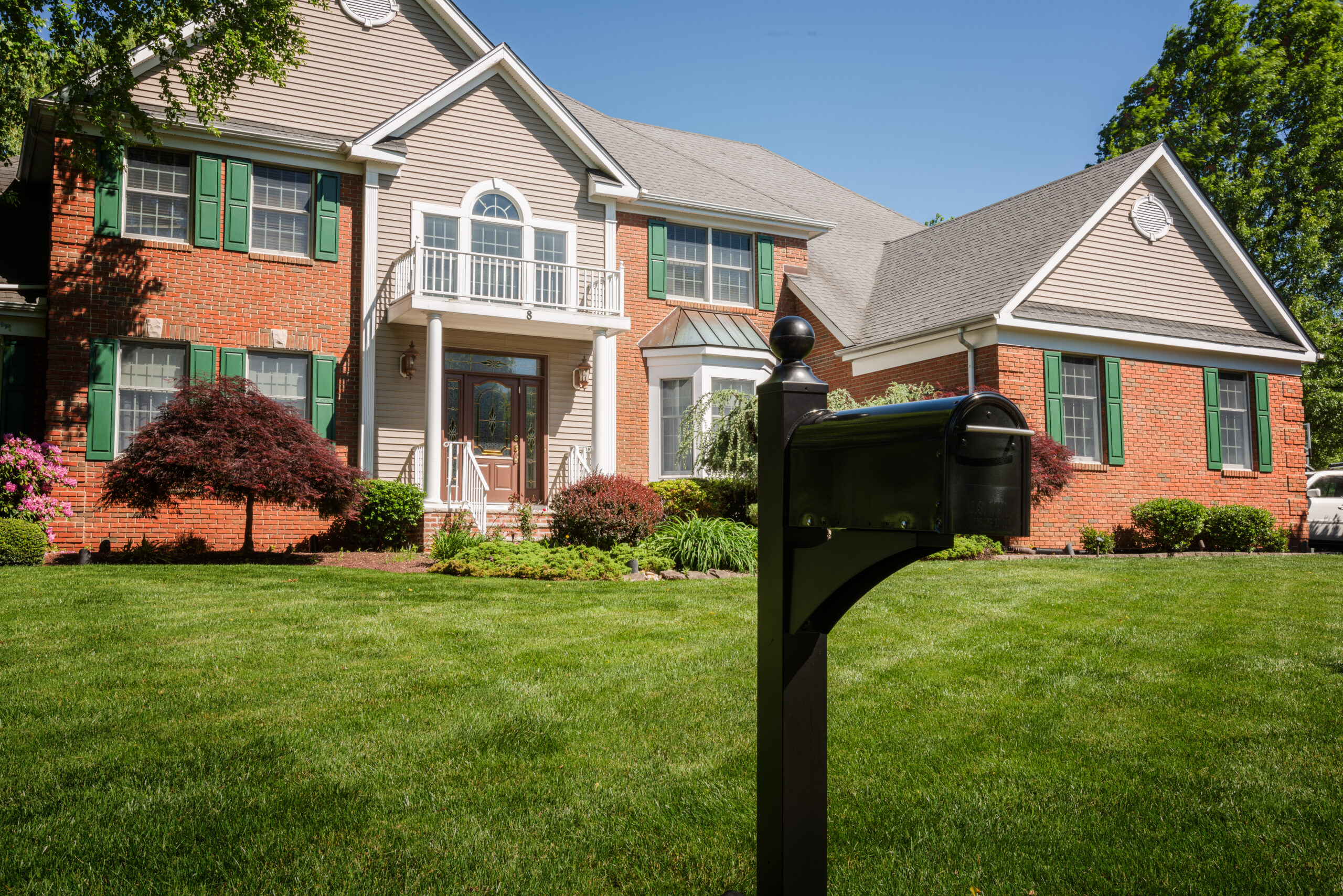 Two-story brick house with green shutters and a manicured lawn, black mailbox in the foreground.