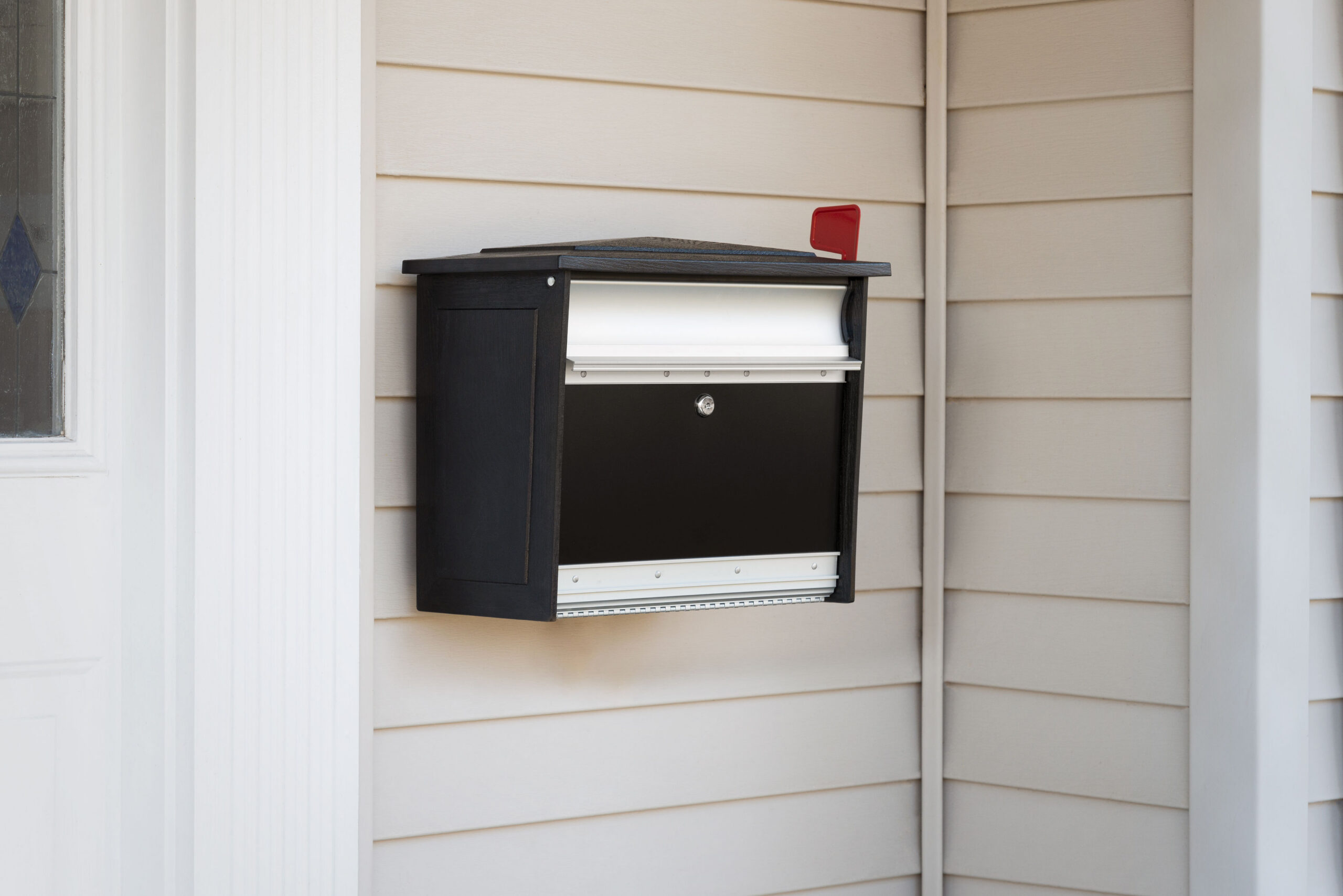 Black wall-mounted mailbox with a red raised flag on beige siding near a white door frame, facing outward from the house.