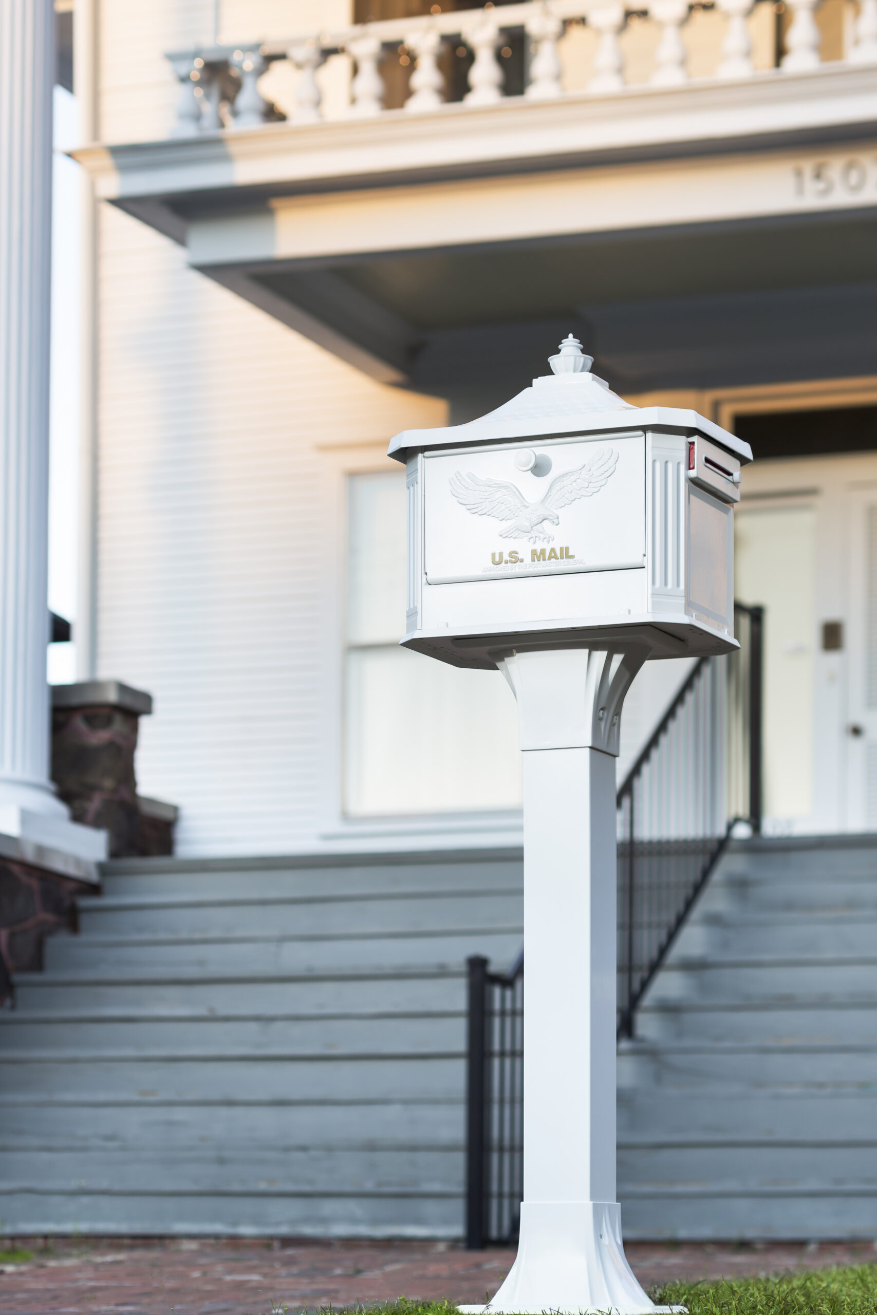 White U.S. Mail mailbox on a tall white post in front of a light-colored house with stairs and railing.