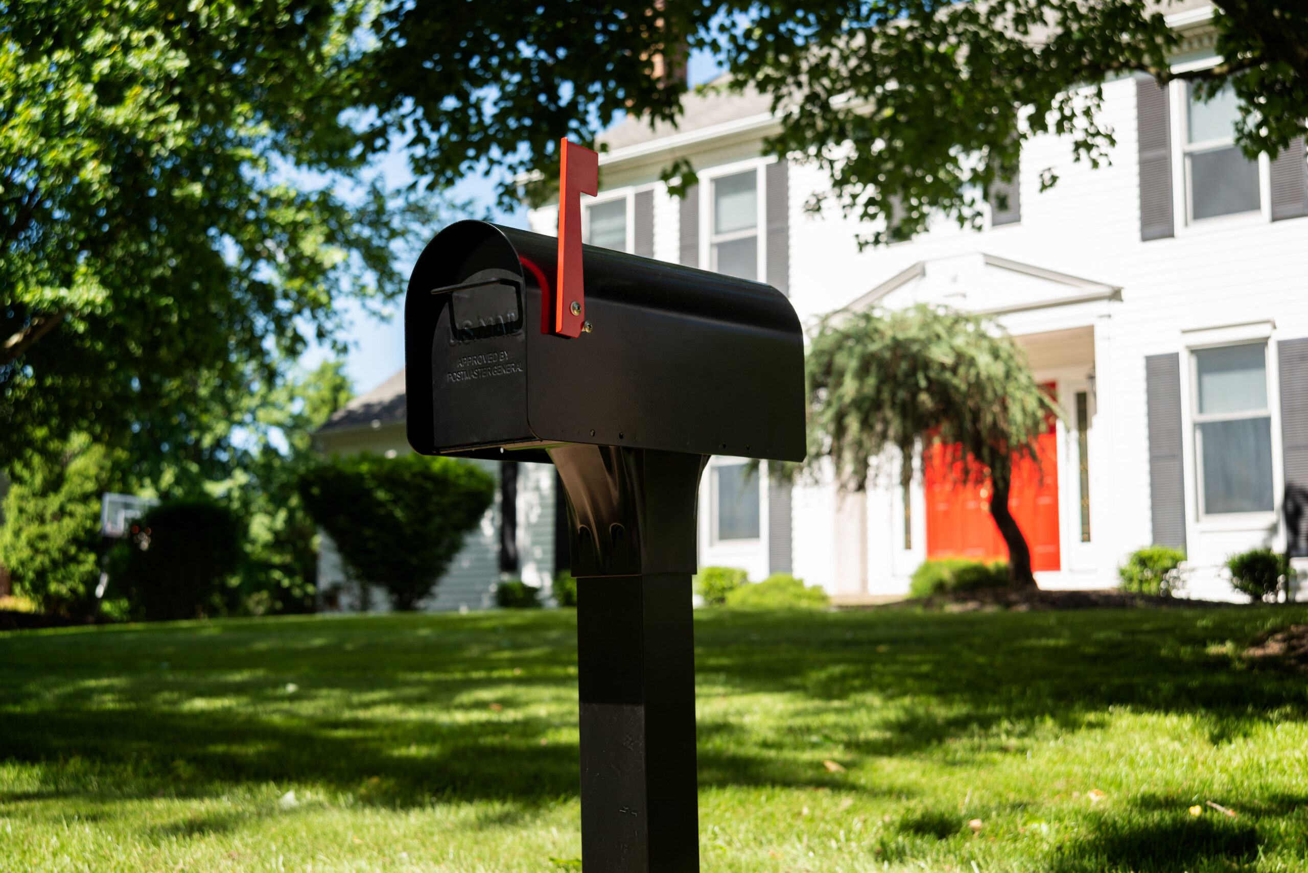 Black curbside mailbox with red flag in a sunny suburban yard, white house with red front door in background