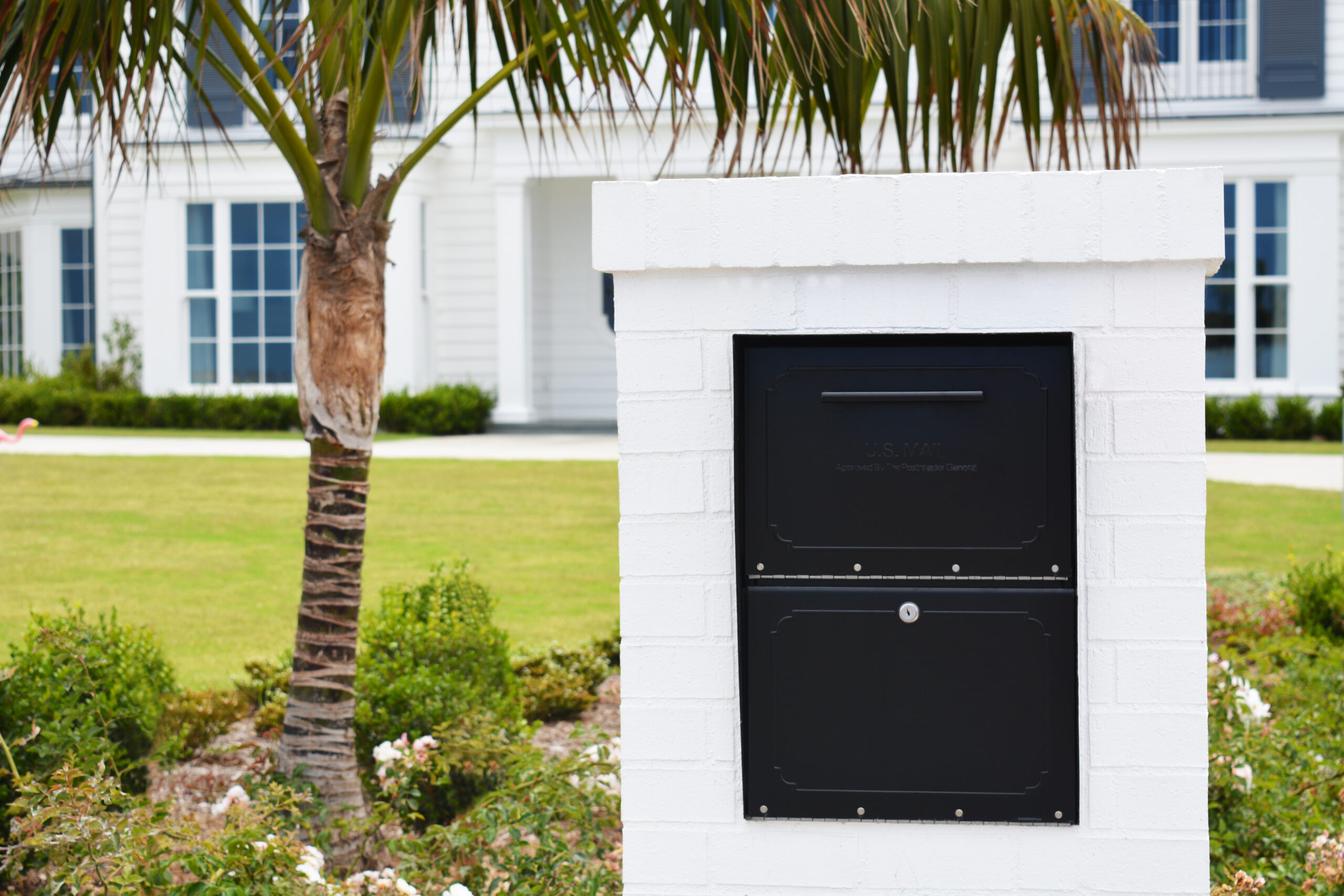 White brick mailbox with a two-compartment black metal door set in front yard of a house, with palm tree and lawn in the background.
