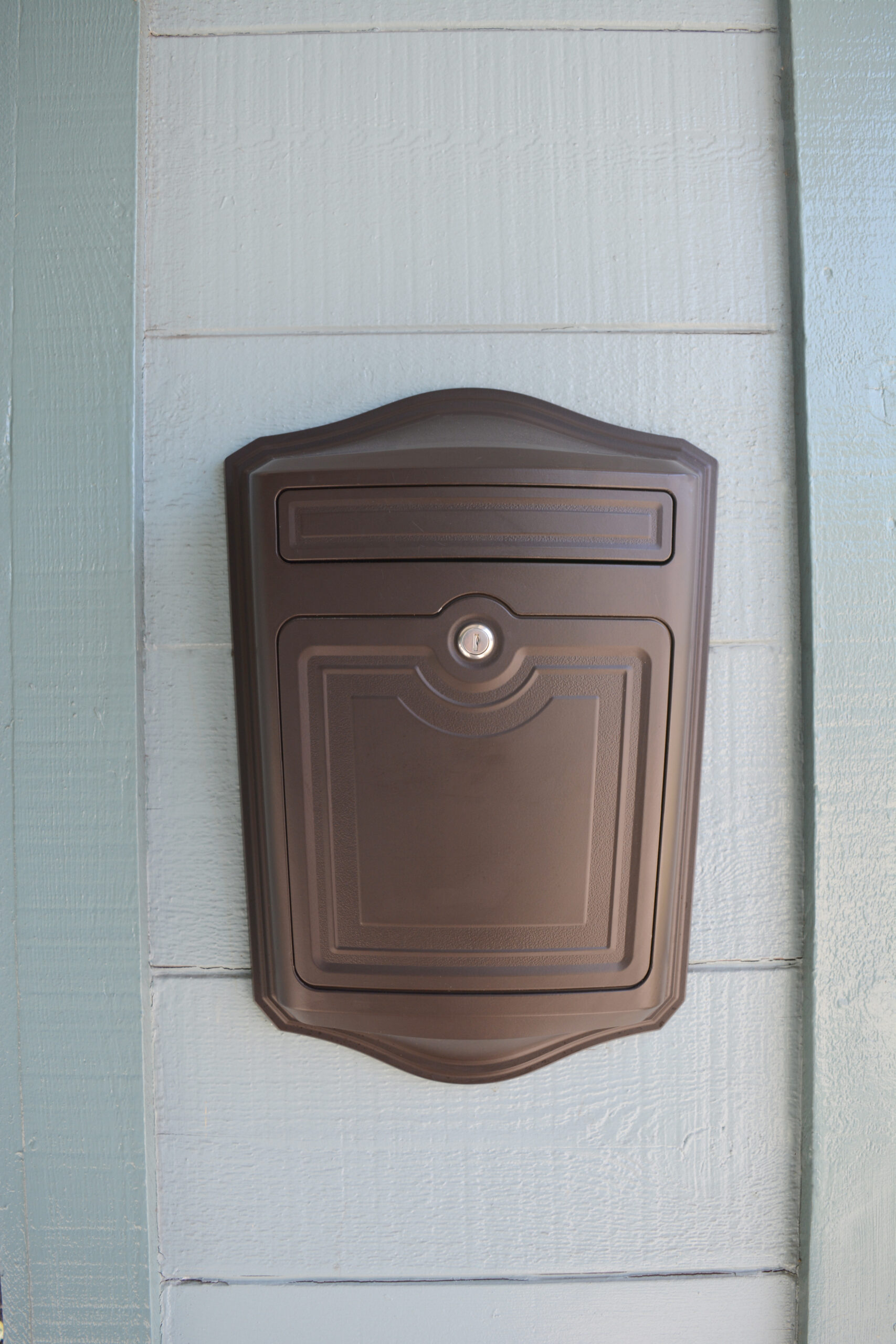 Brown metal mailbox with a rounded top mounted on pale blue wooden siding, including a front door and mail slot area with a small lock above the panel.