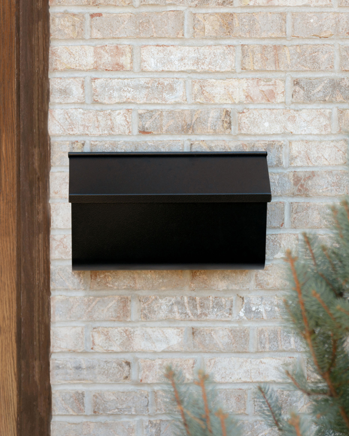 Black wall-mounted mailbox attached to a light brick exterior wall, with a wooden beam on the left and some greenery in the foreground
