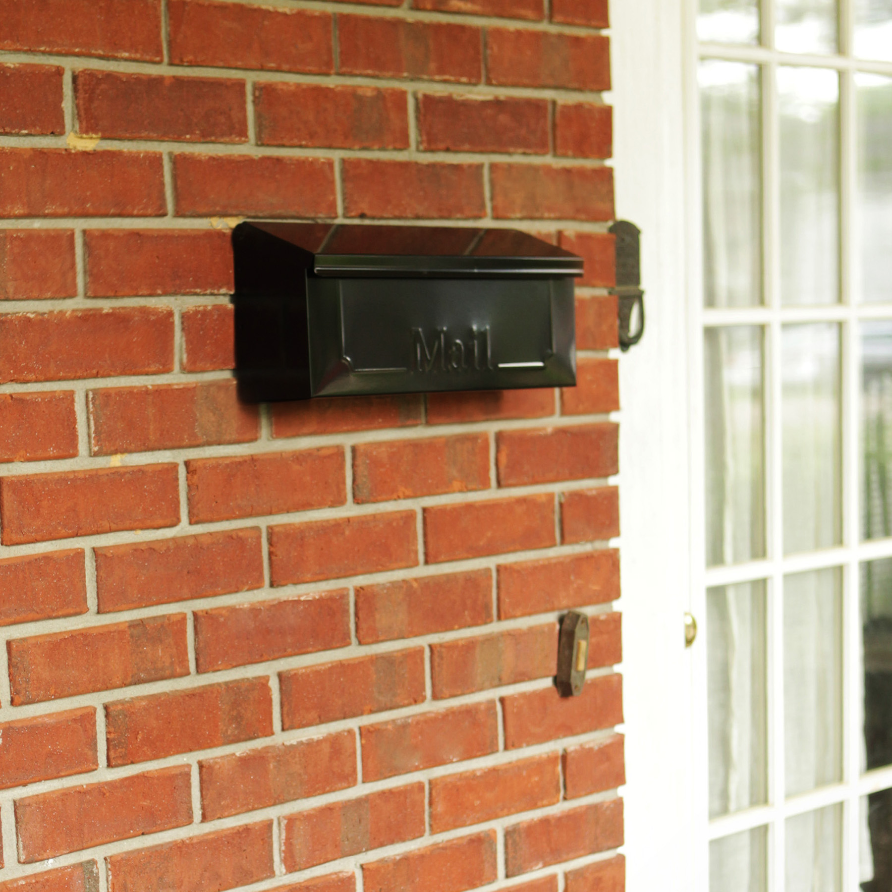 Black wall-mounted mailbox on a red brick exterior beside a white-framed door window area.