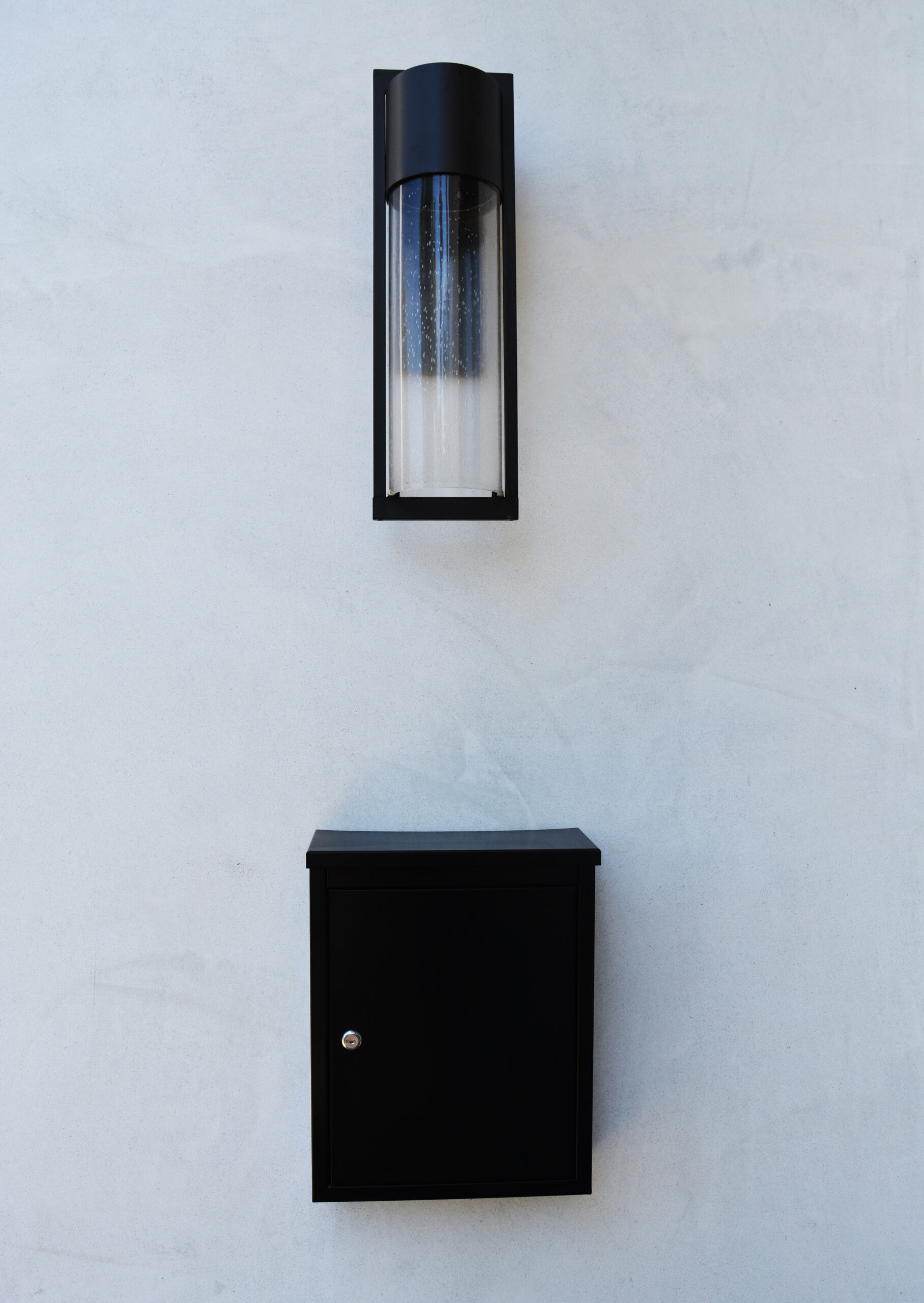 Black rectangular outdoor mailbox mounted on a light gray stucco wall with a tall black cylindrical wall light above it.