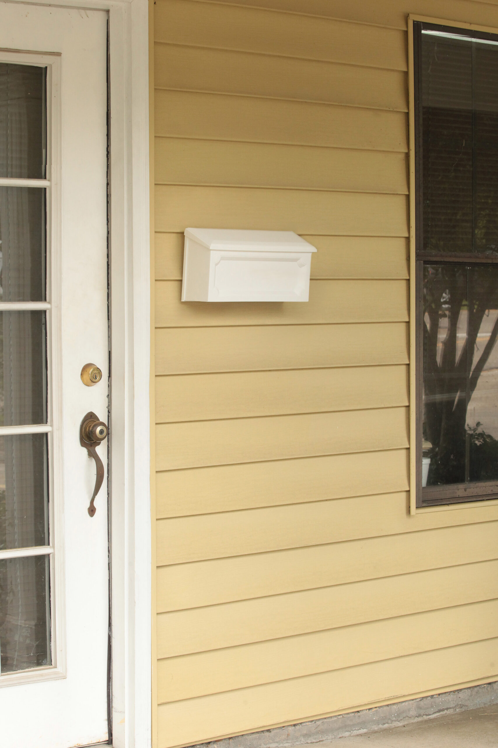 White wall-mounted mailbox on yellow siding beside a glass-panel door and a window.