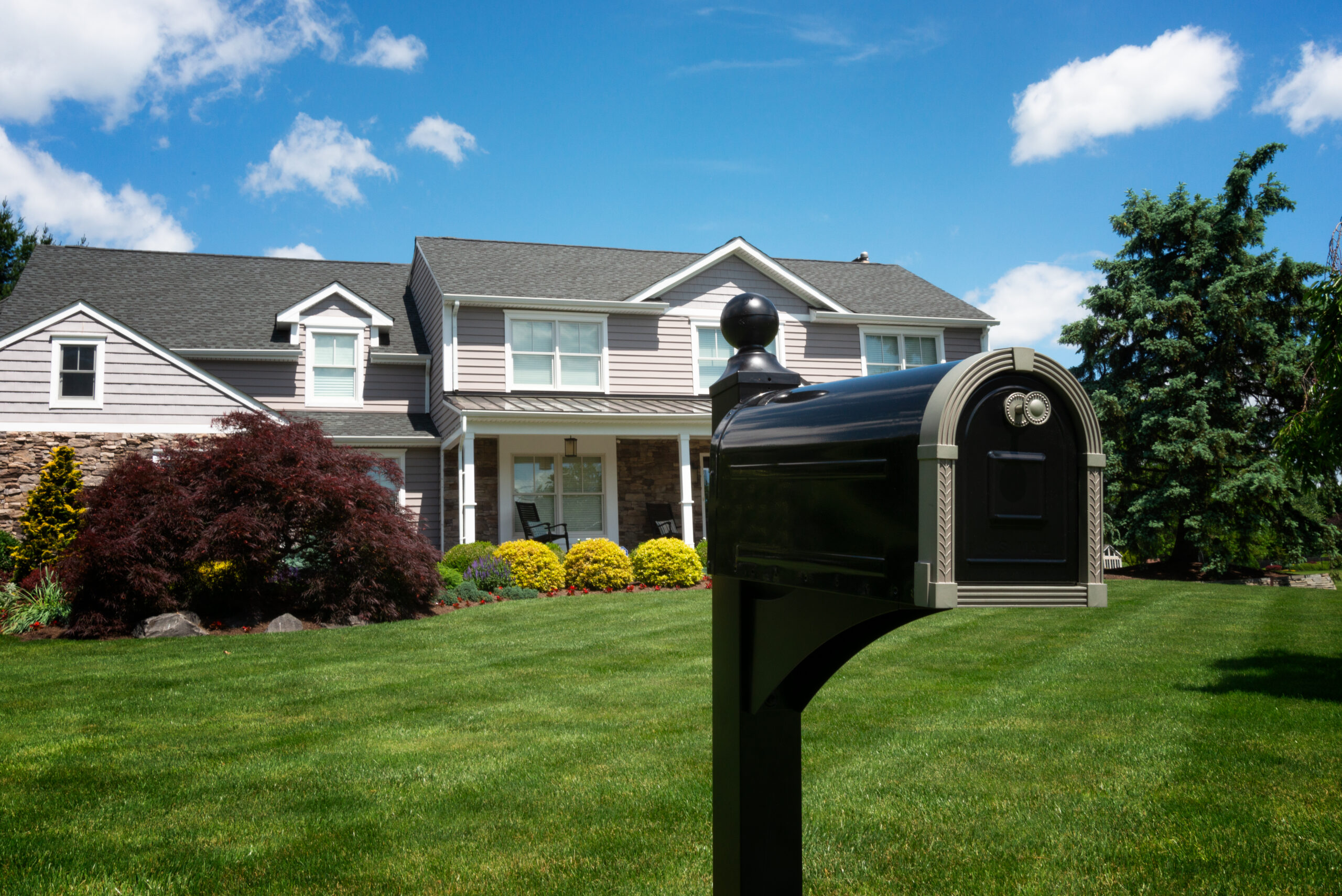 Two-story gray suburban house with a manicured lawn and colorful shrubs, seen from a yard with a prominent black curved mailbox in the foreground under a bright blue sky.