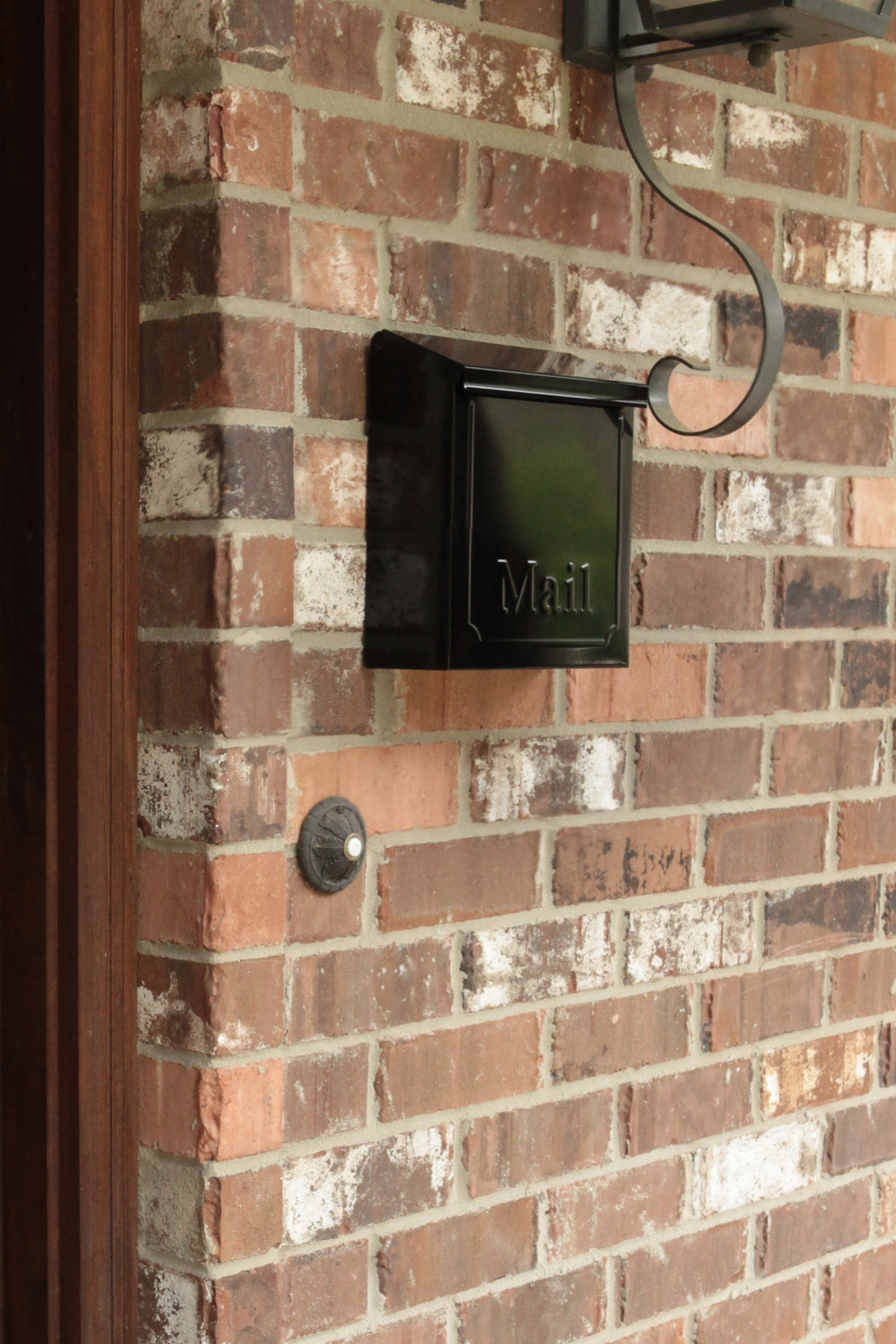 Black wall-mounted mailbox with 'Mail' embossed, attached to a brick exterior wall under a decorative curved metal bracket.
