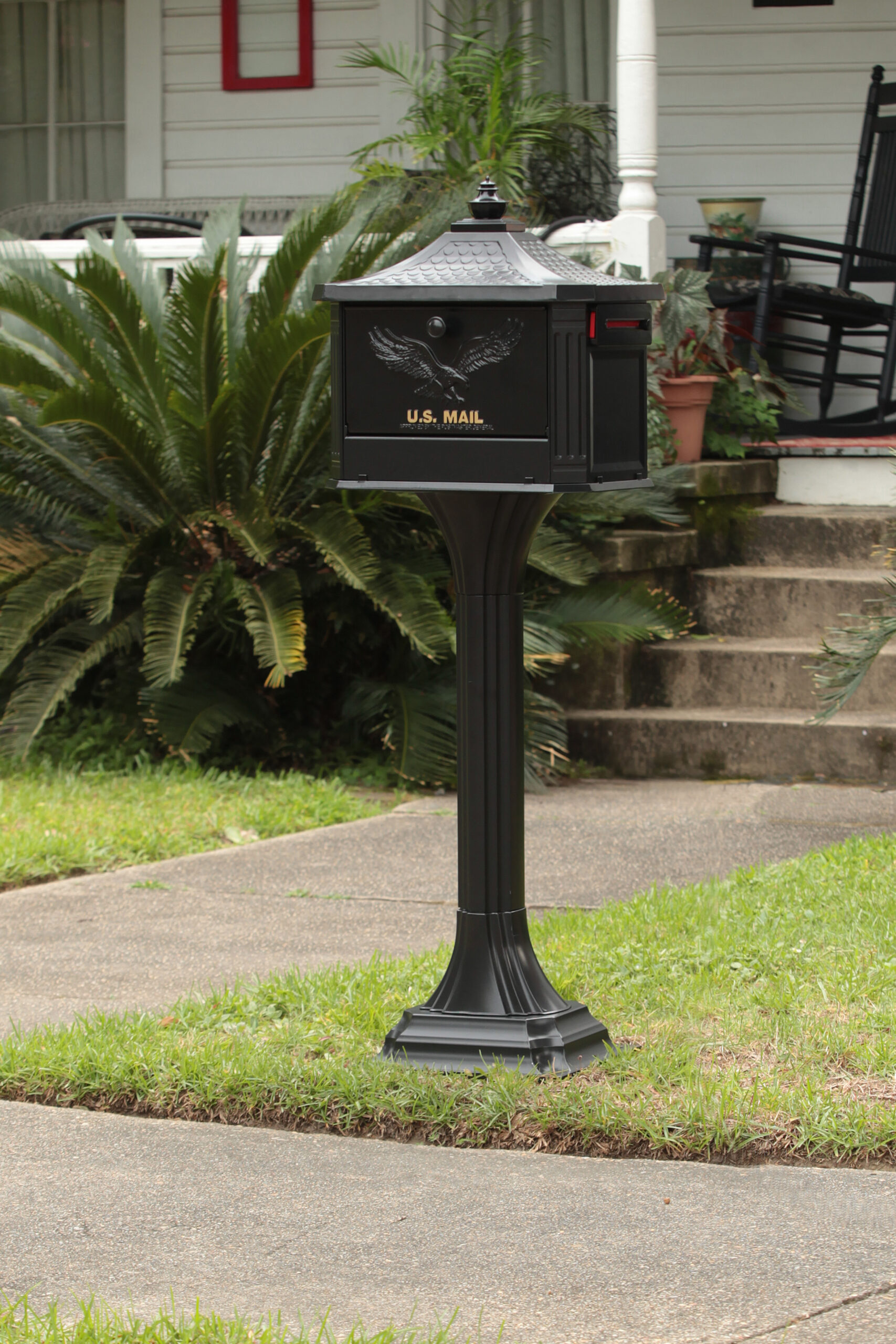 Black U.S. Mail mailbox on a tall pedestal in a suburban front yard, with greenery and a porch in the background.