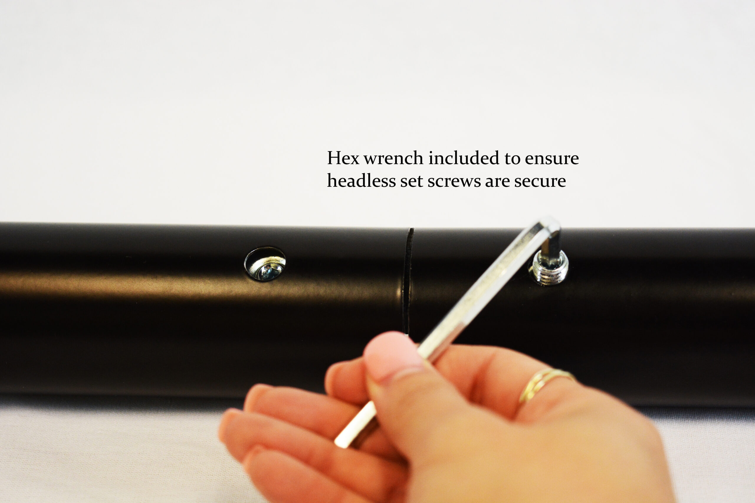 Hand holds a silver hex wrench to tighten a headless set screw on a black cylindrical rod; caption reads 'Hex wrench included to ensure headless set screws are secure'.