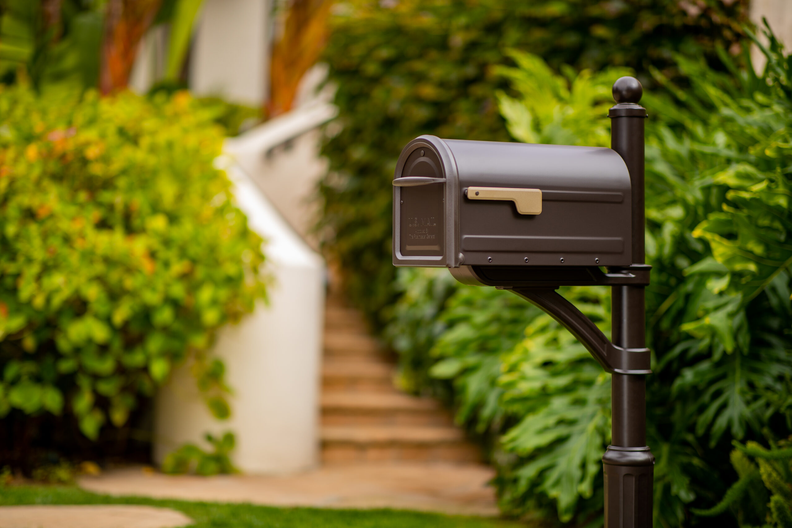Gray outdoor mailbox on a dark post in a landscaped front yard with green shrubs and steps in the background