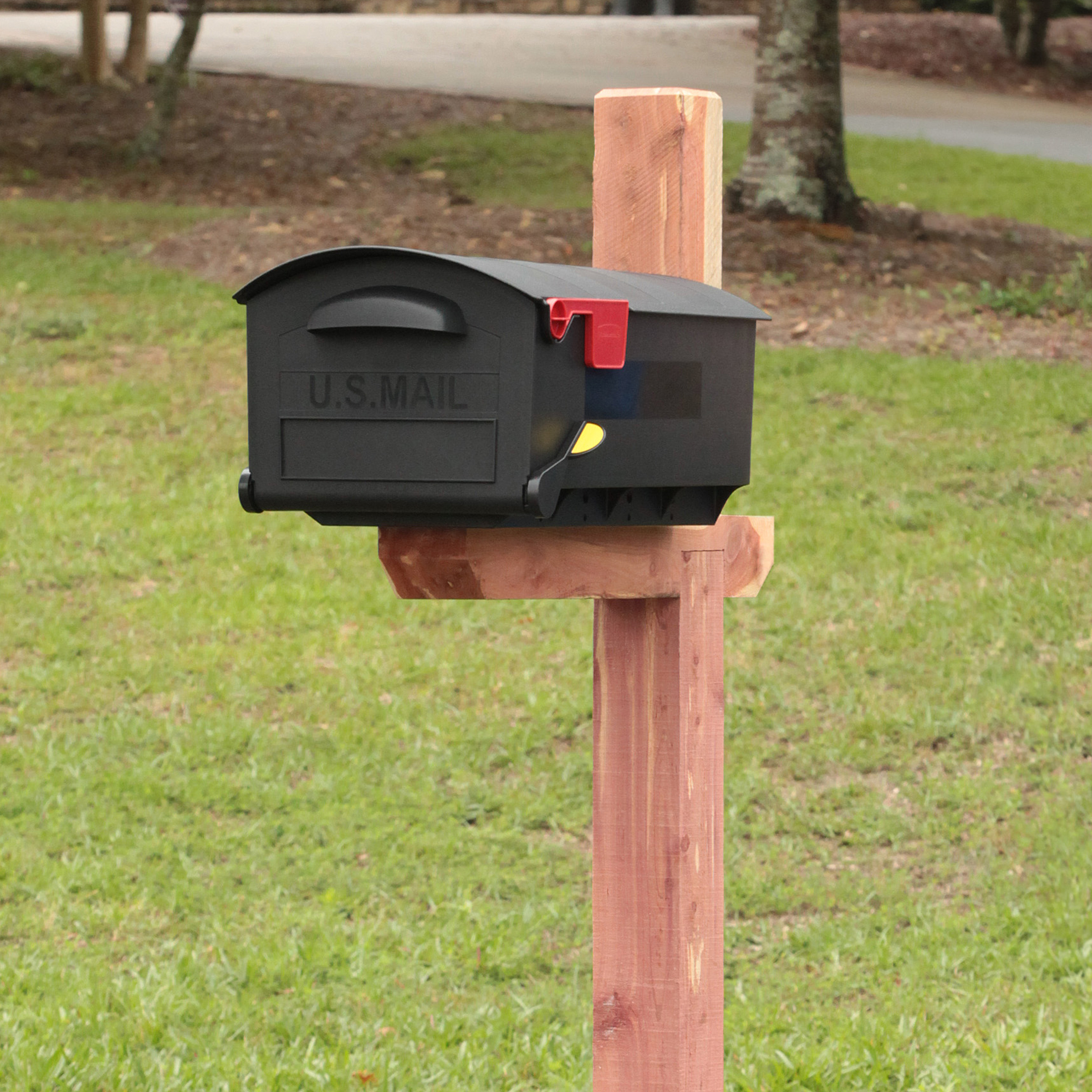 Black U.S. Mail mailbox with a red flag raised, mounted on a wooden post in a grassy yard.