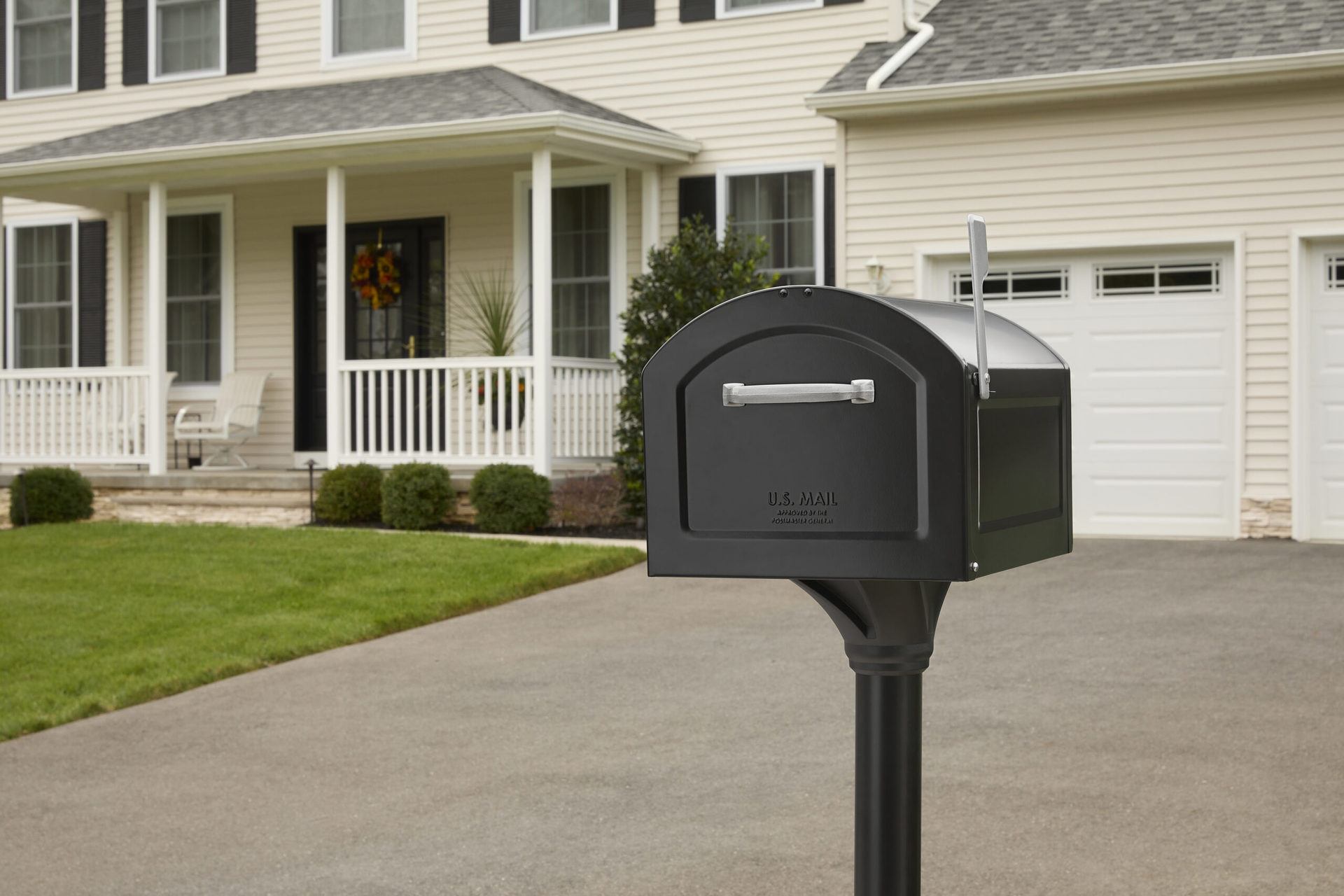 Mailbox on a black post in a suburban driveway with a two-story beige house and front porch in the background