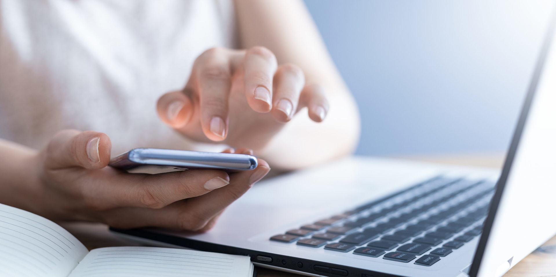 Person using a smartphone while a laptop and open notebook sit on a desk side by side