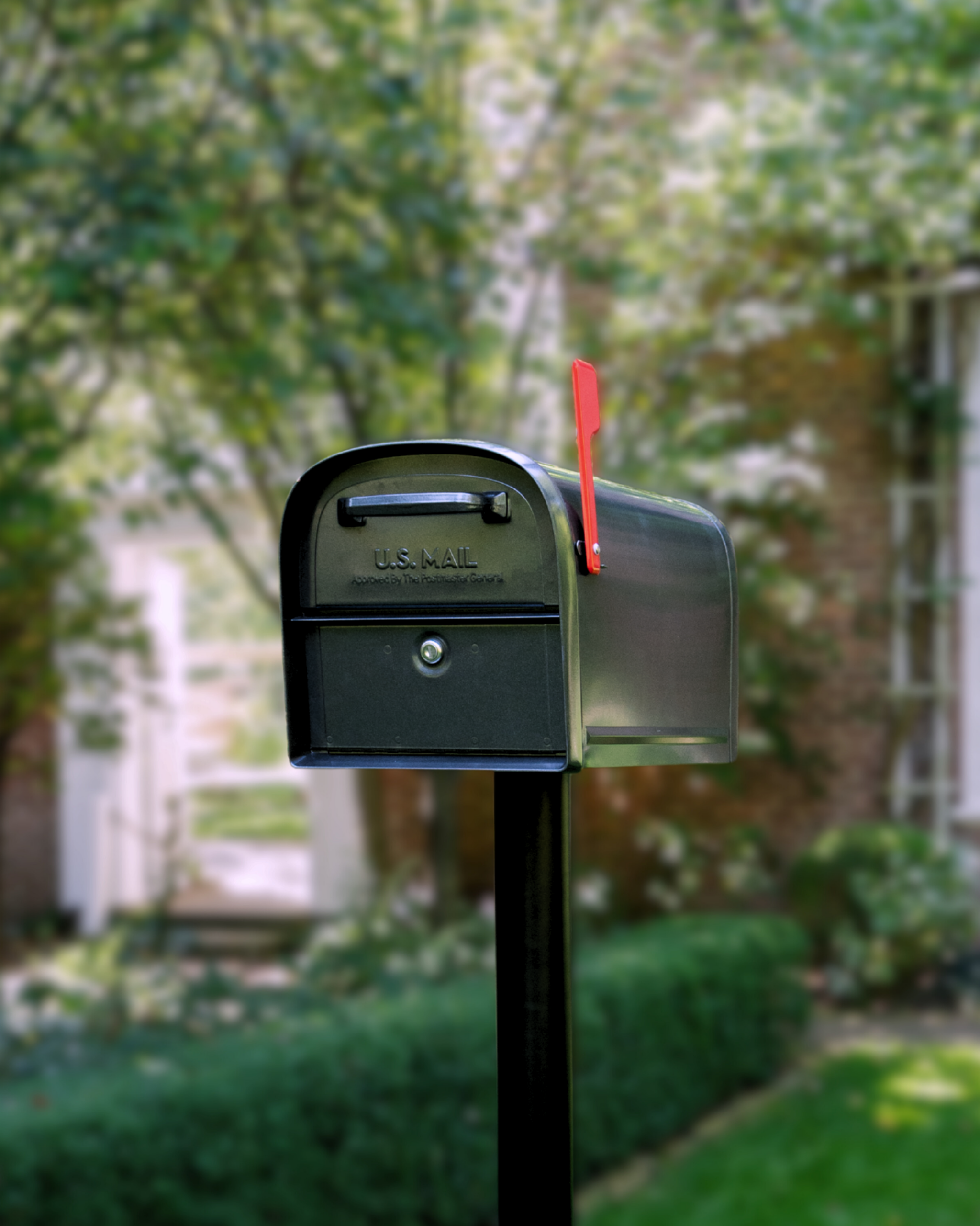 Green U.S. Mail mailbox on a post with the red flag raised, set against a blurred suburban yard backdrop.
