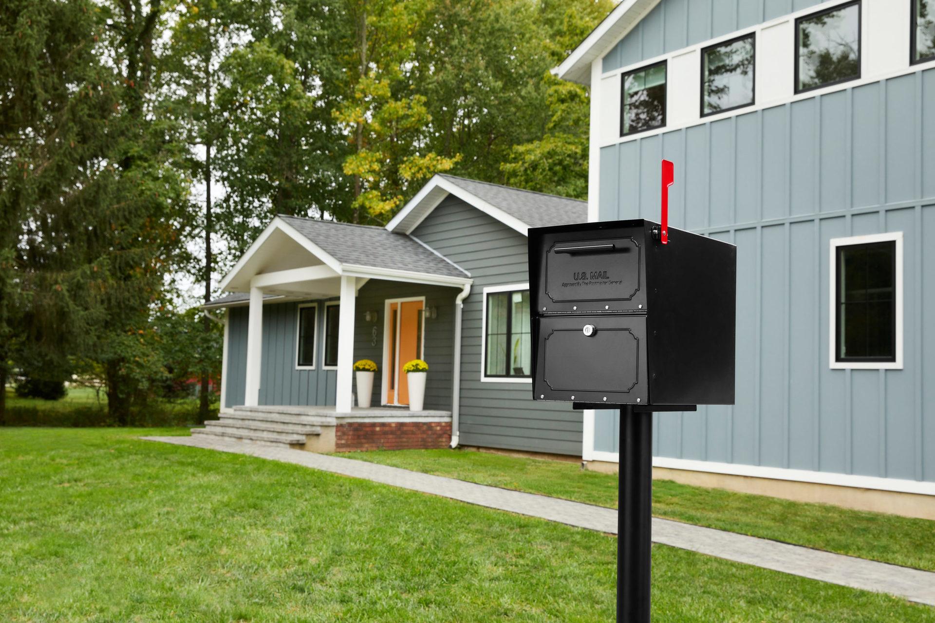 Black mailbox on a post with a red flag in the front yard of a gray house with white trim and a front porch. The mailbox reads