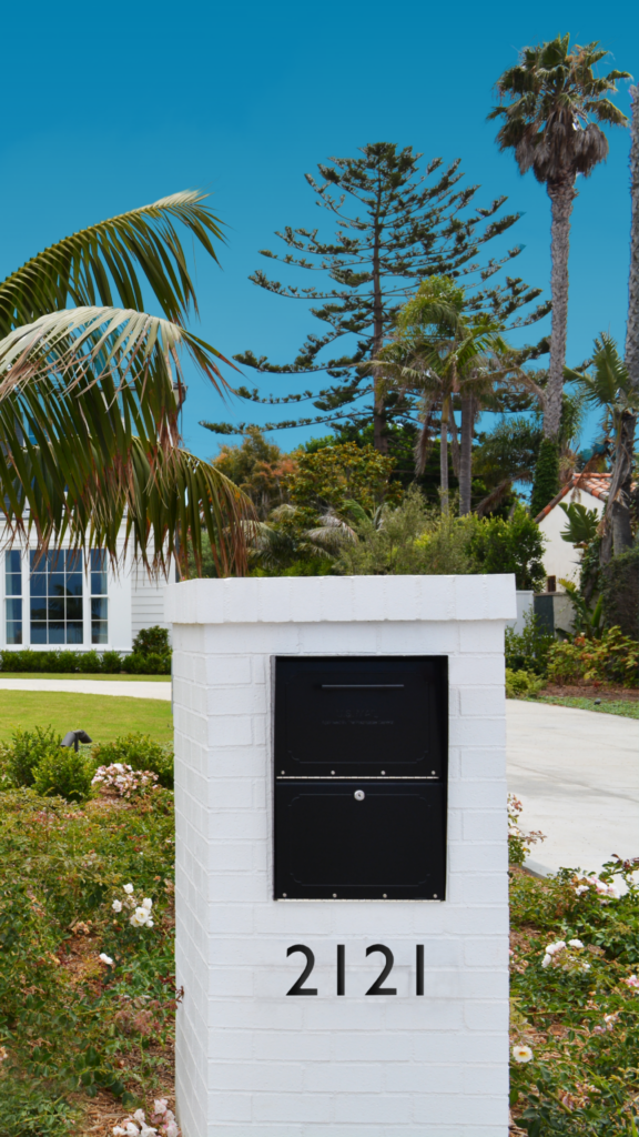 White brick mailbox with a black metal door and the address 2121, in a landscaped yard with palm trees and a bright blue sky.