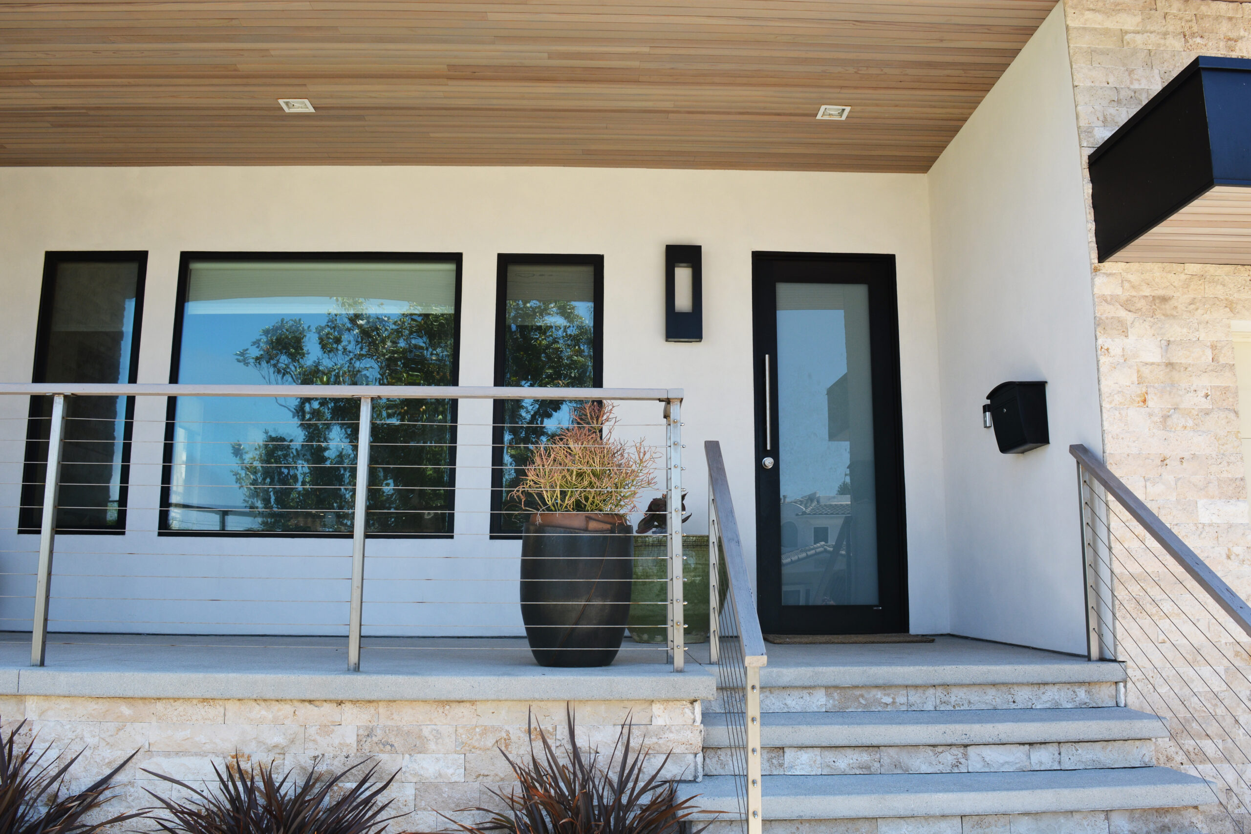 Modern front porch of a contemporary house with black-framed glass door, three tall windows, metal railing, and a large potted plant on the deck.