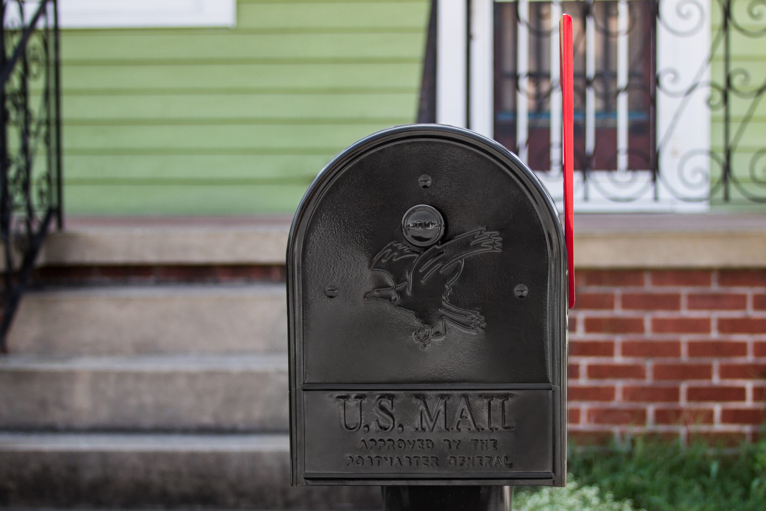 Black US Mail mailbox with a raised eagle emblem and an upright red flag in front of a green house.