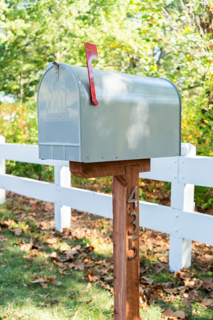Silver metal U.S. mailbox with a red flag, perched on a wooden post marked with the number 435 beside a white fence in a leafy yard.