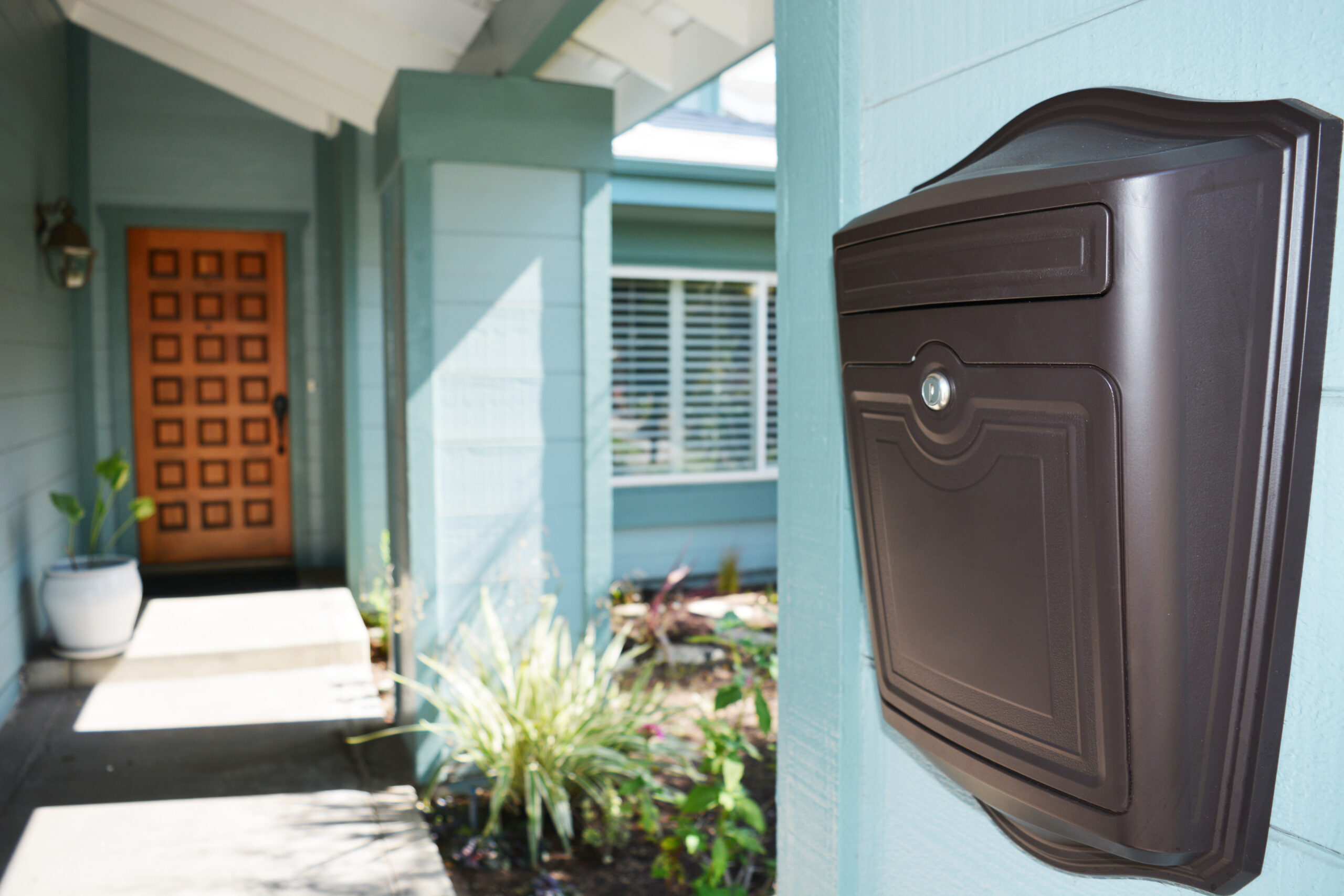 Black wall-mounted mailbox on a blue house, with a wooden front door and a small garden path nearby.