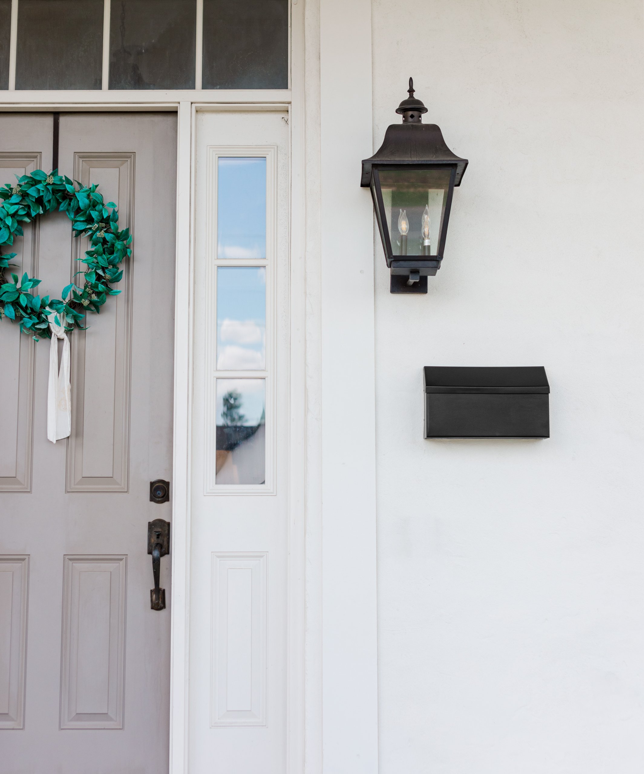 Beige front door with decorative panels, a green wreath on the left, glass sidelights, and a black outdoor lantern on the right.