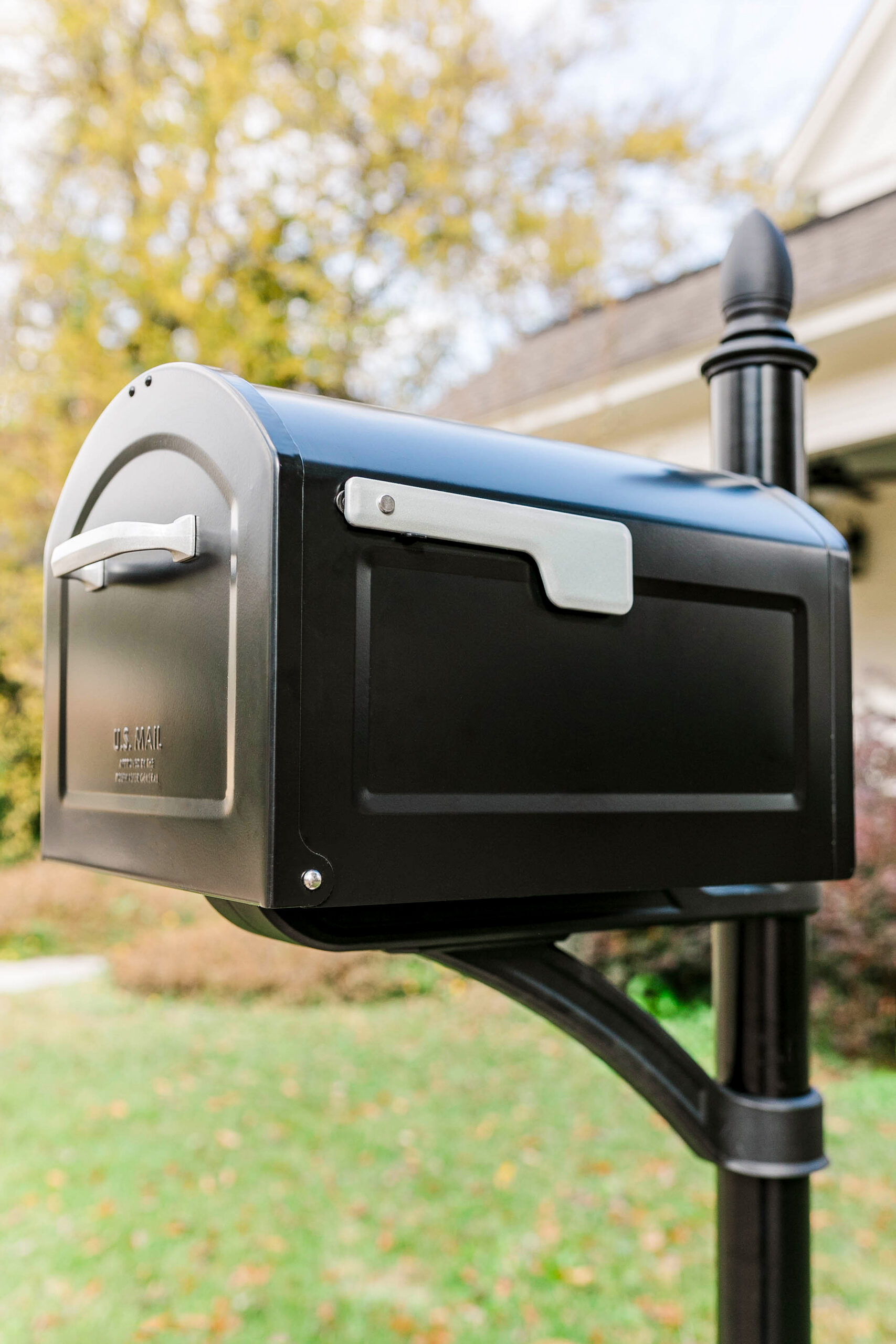 Black metal mailbox on a post in a yard with autumn trees in the background, suburban setting.