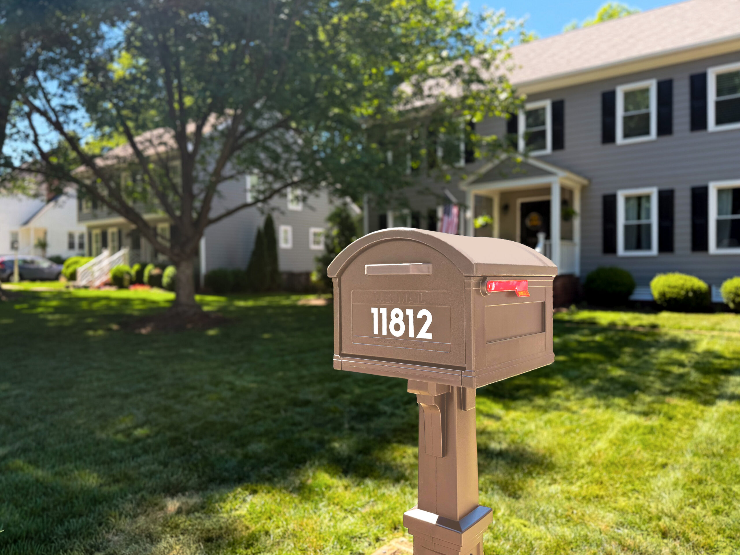 Brown residential mailbox with a red flag raised in a suburban yard, houses and trees in the background on a sunny day.