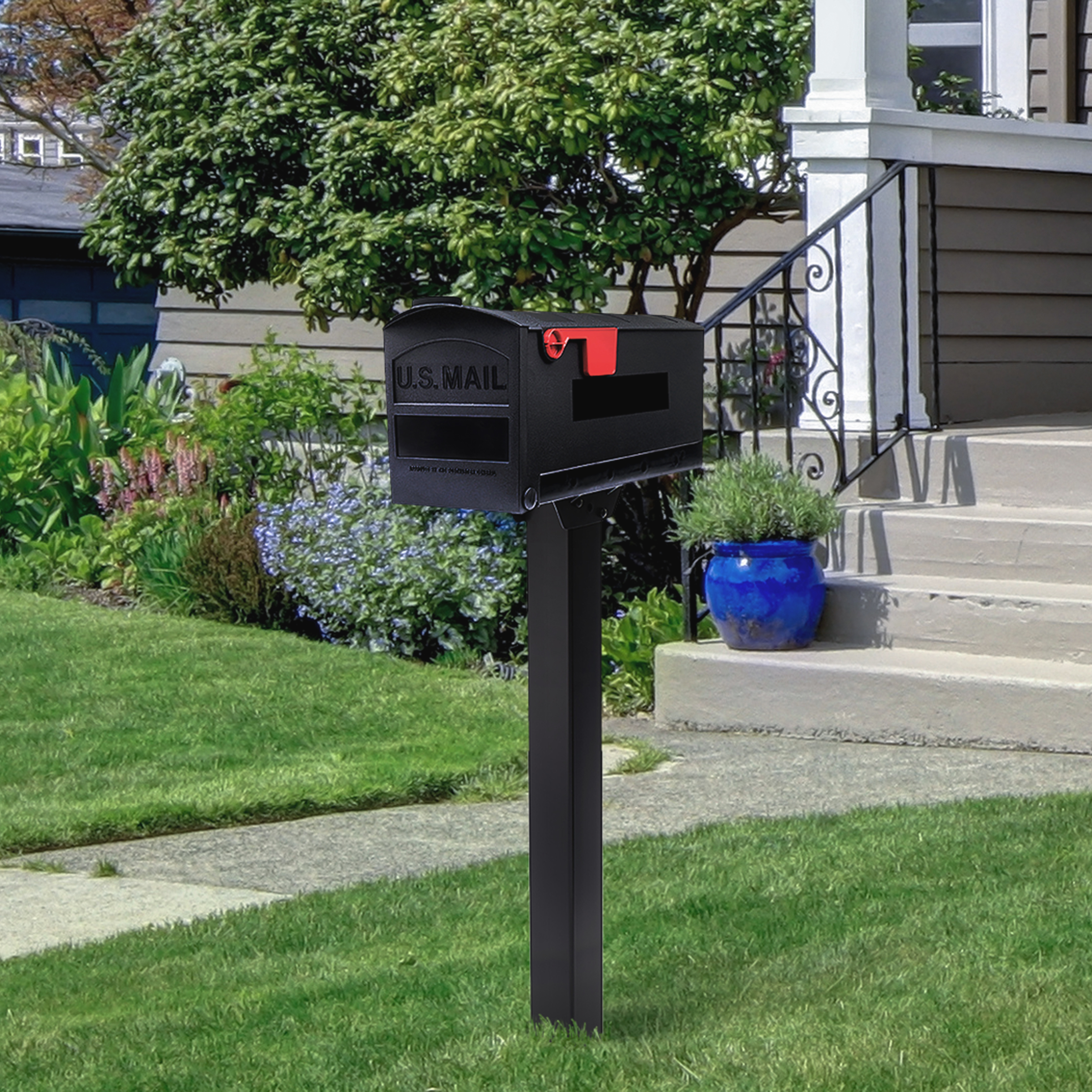 Black curbside USPS mailbox with red flag raised, mounted on a post in a front-yard lawn near a house with steps and a blue planter.