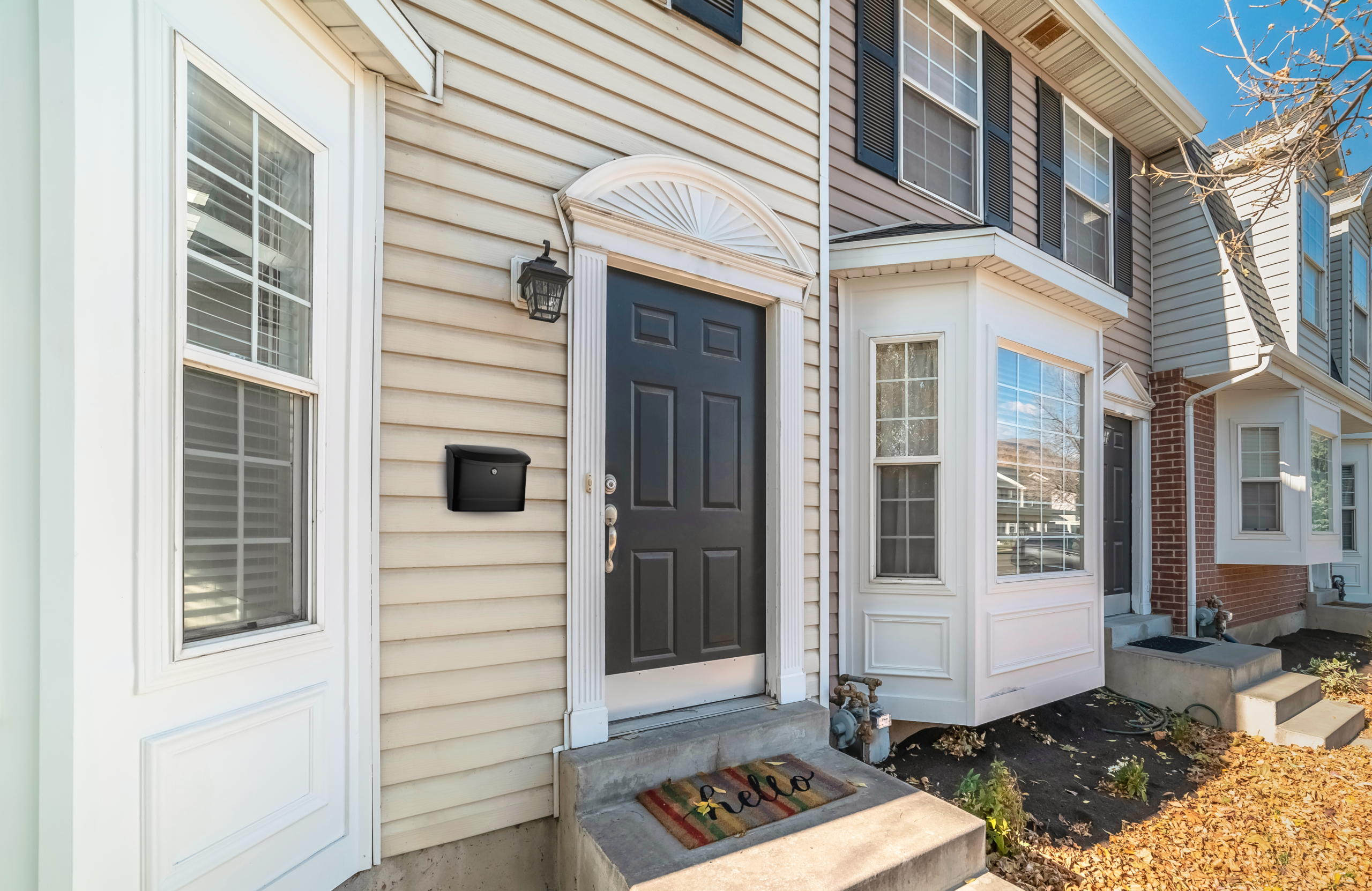 Row of beige townhouses with a black front door, arched fanlight above, white trim, a lantern light, and a welcome mat on concrete steps near a mailbox.