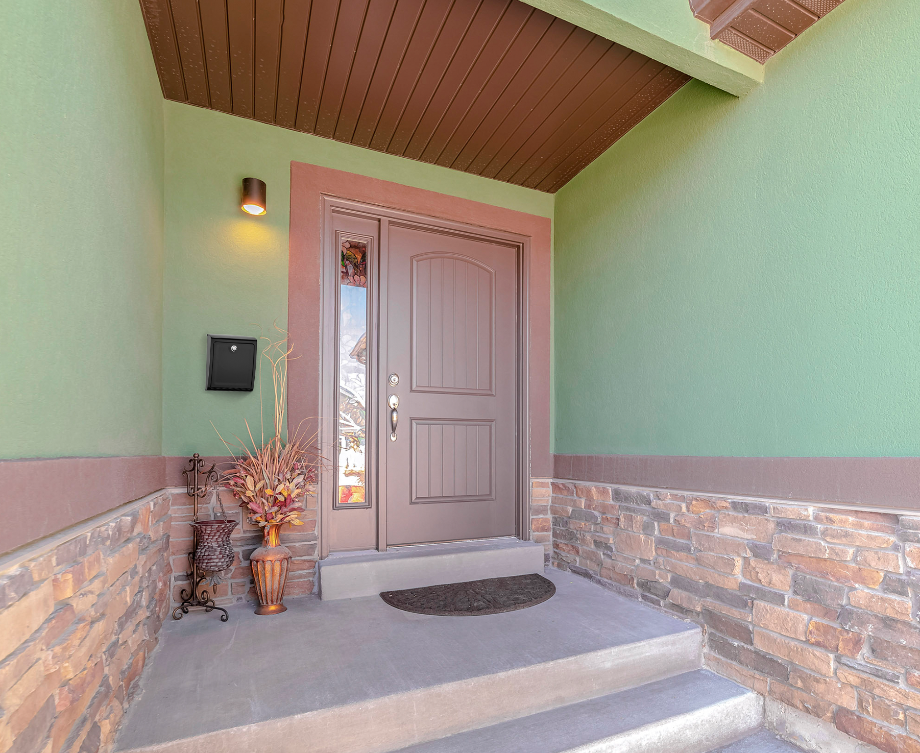 Front entryway with a brown paneled door, green exterior walls, and stone veneer along the bottom edge; a black mailbox is mounted on the left wall beside a decorative plant.